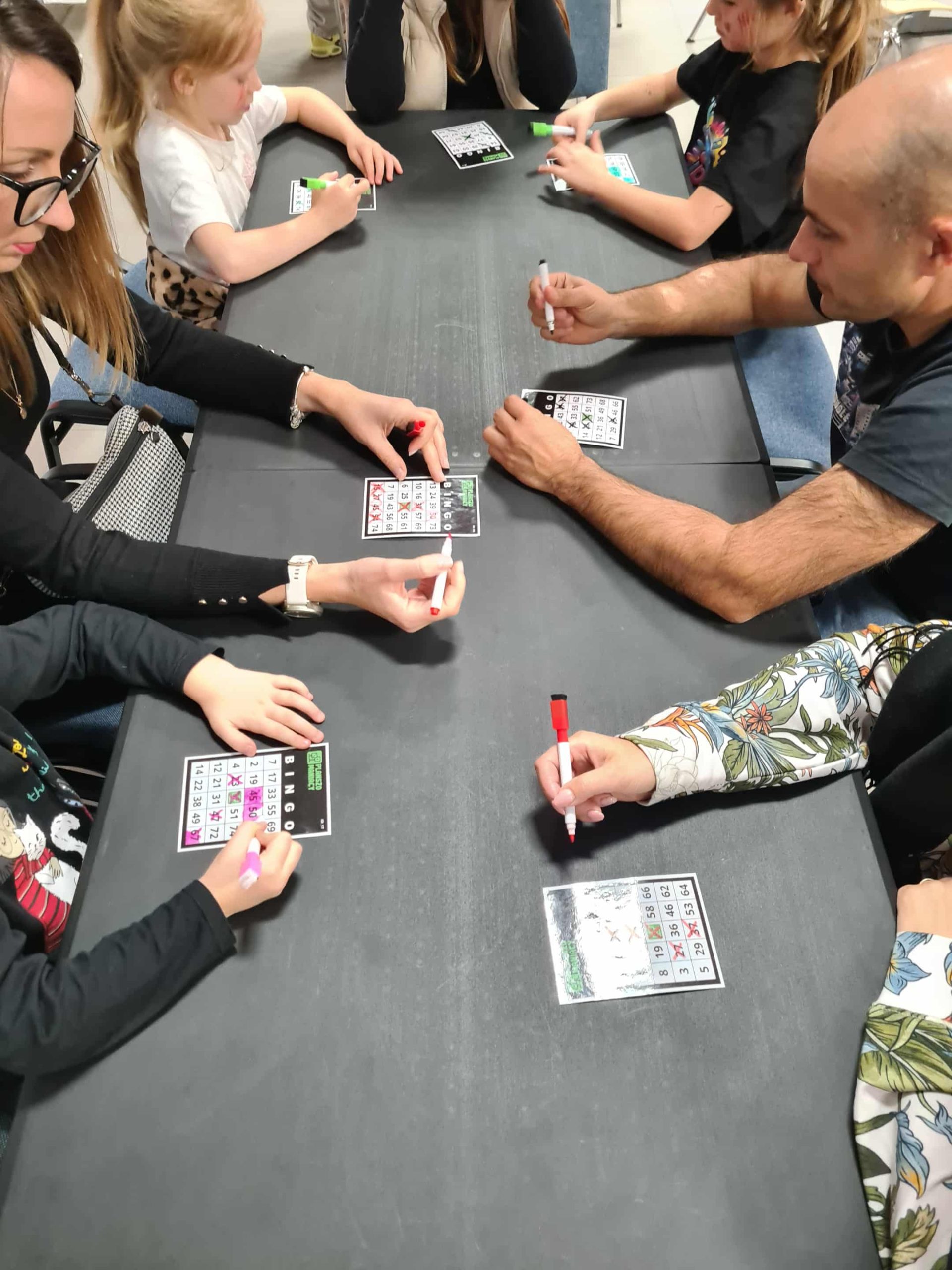 Rodziny grają w bingo podczas planszówkowej nocy w bibliotece, skupieni nad kartami i kolorowymi markerami.