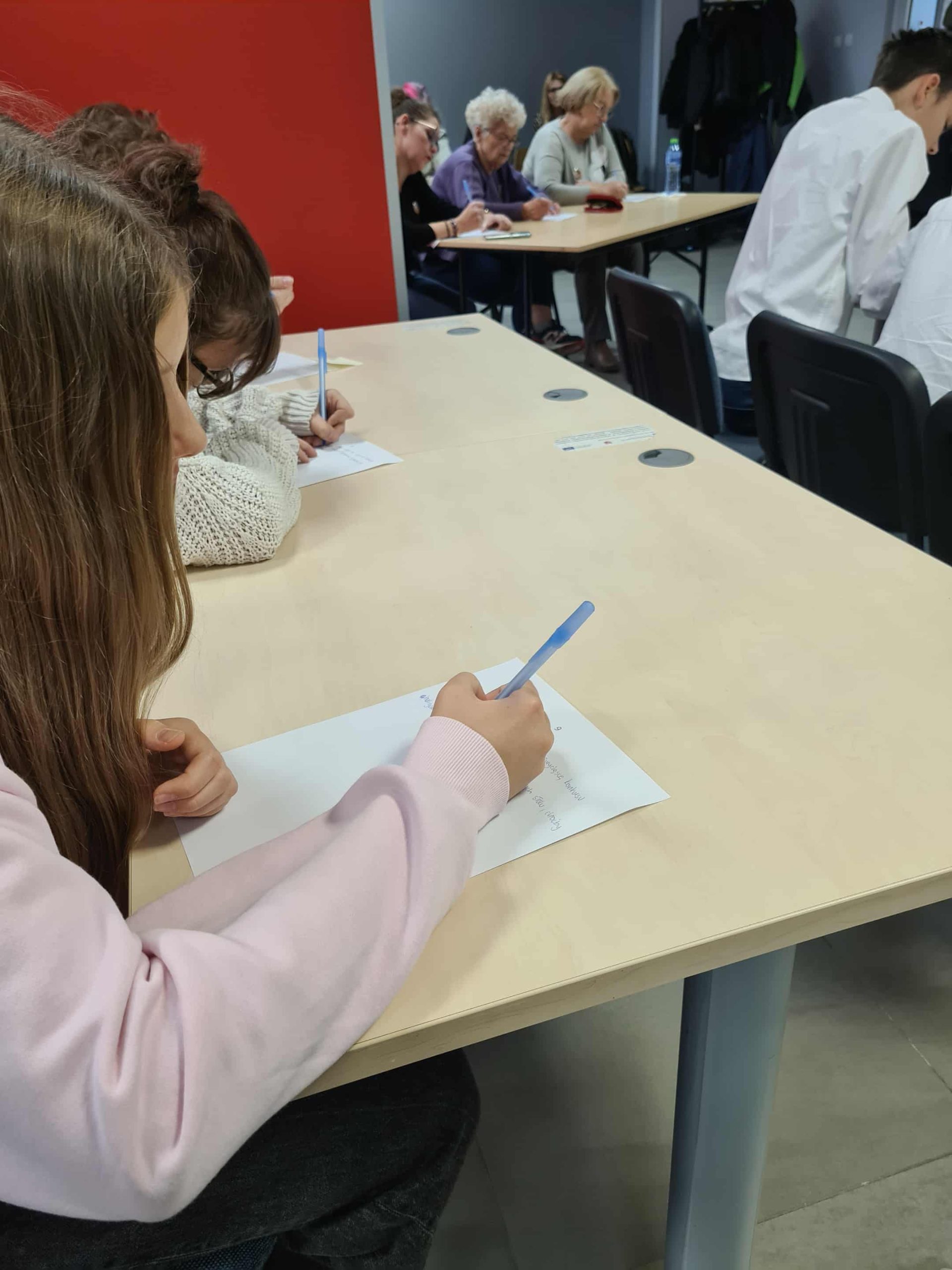 Uczestnicy w różnym wieku piszą dyktando przy stołach w sali biblioteki.