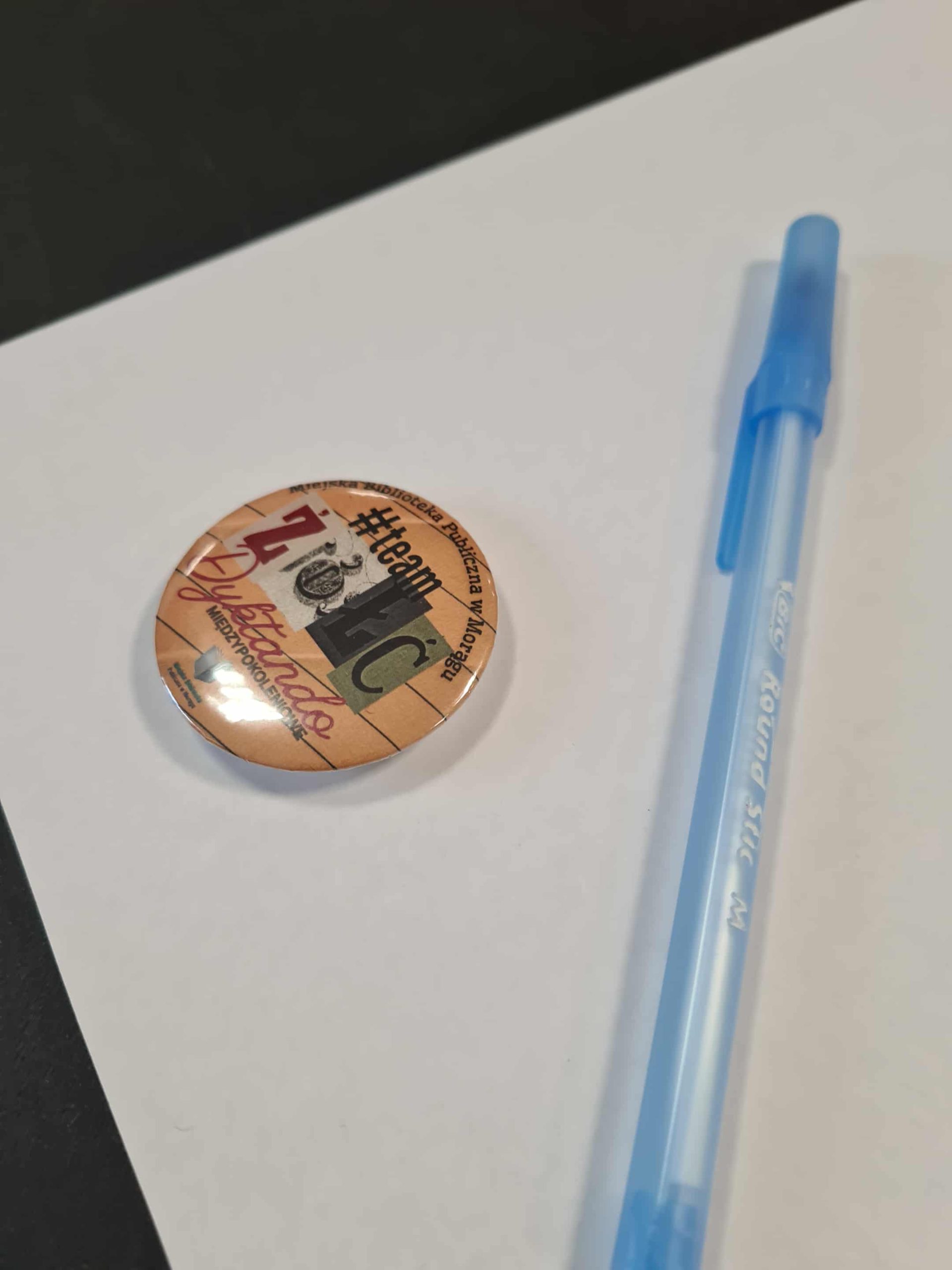Długopis i przypinka z logo Dyktanda Międzypokoleniowego na biurku uczestnika.