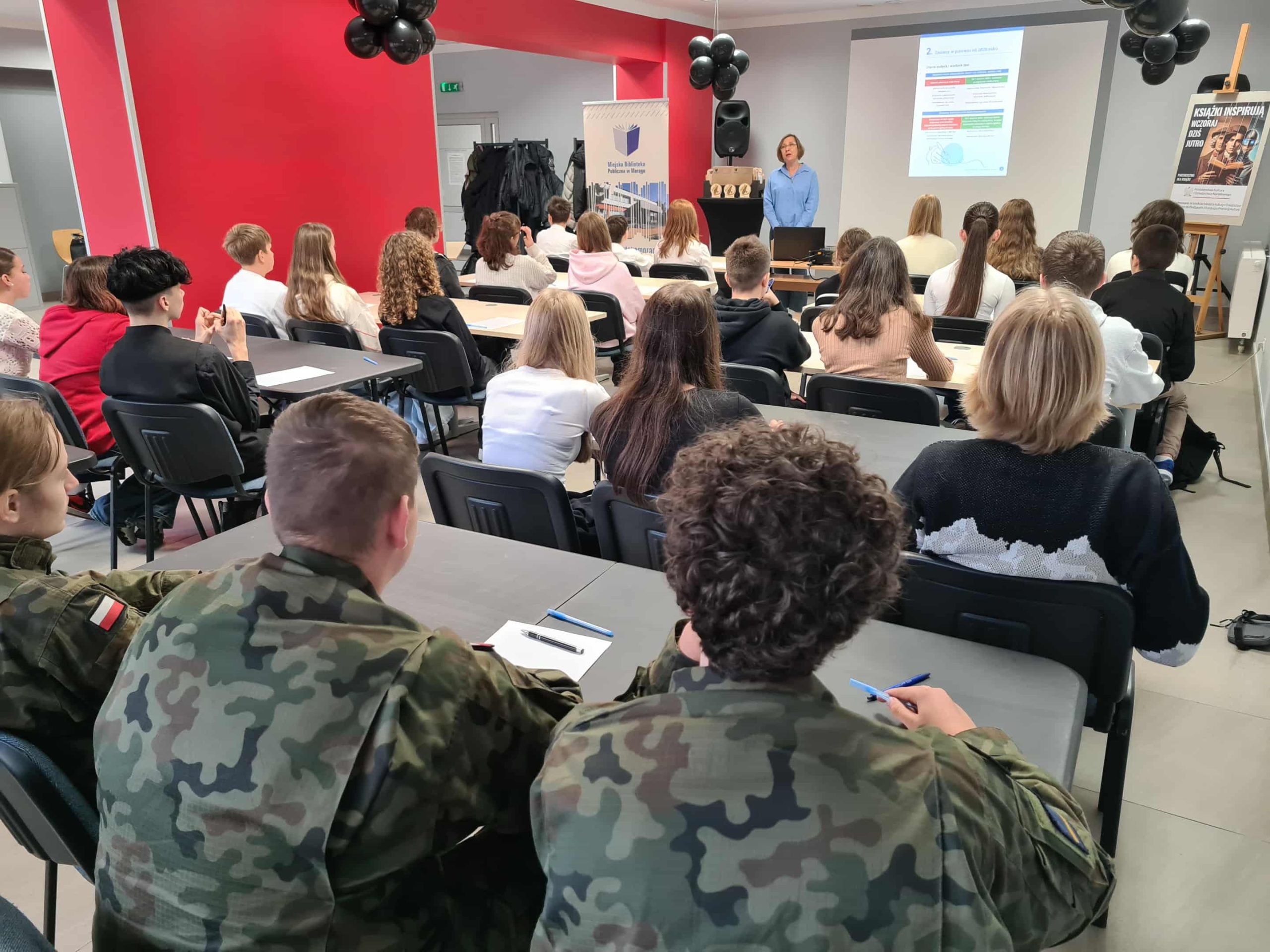 Uczestnicy, w tym młodzież w mundurach, piszą dyktando w czytelni biblioteki.