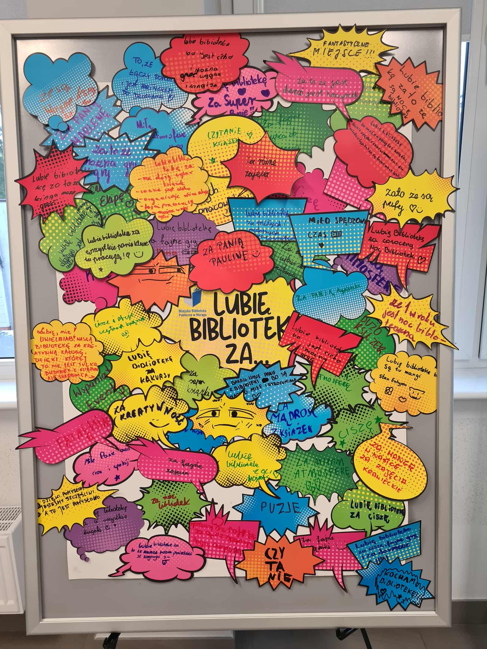 Cała plansza „Lubię bibliotekę za...” w komiksowym stylu, wypełniona dziesiątkami kolorowych chmurek z wpisami czytelników.