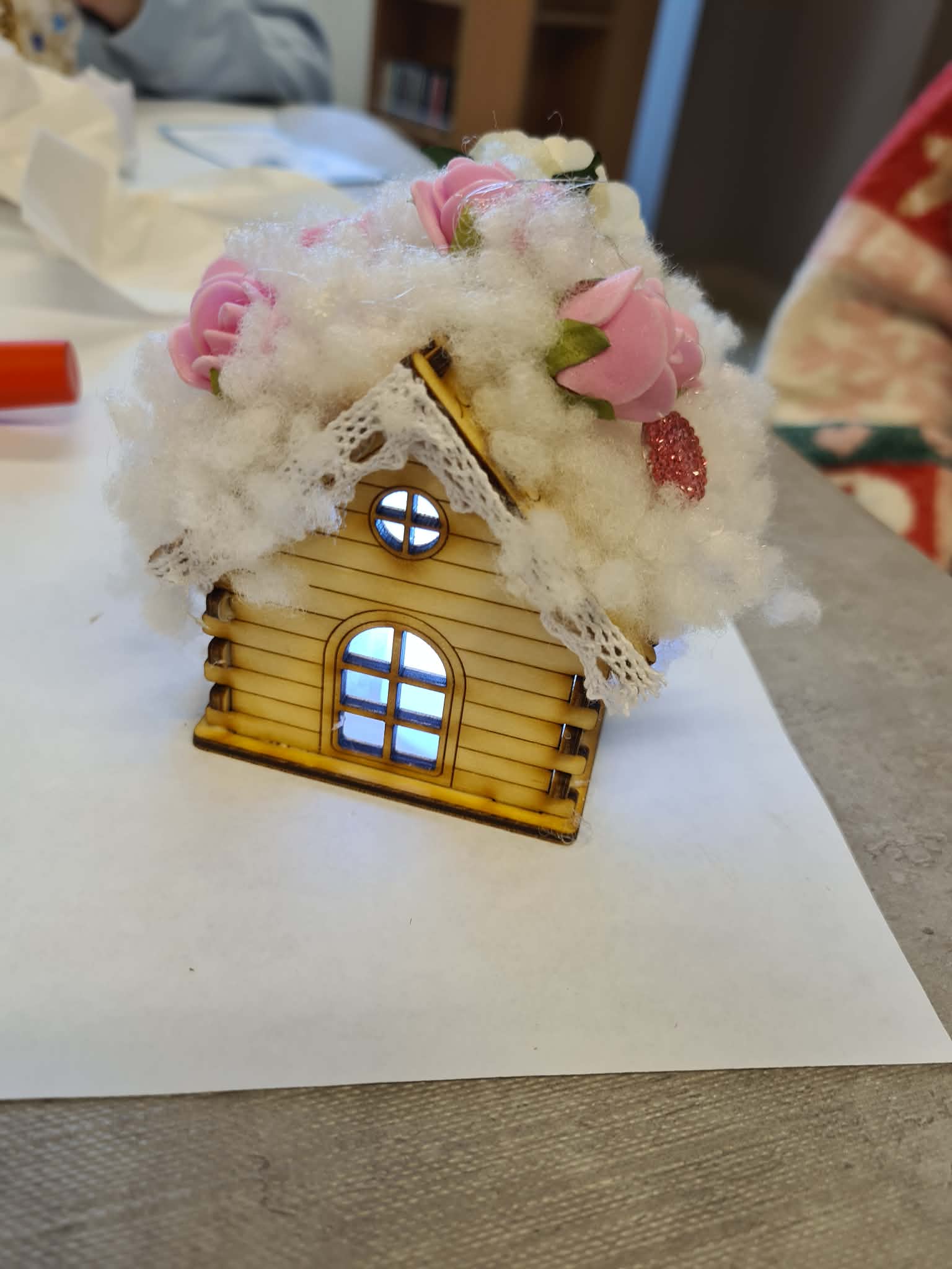 Mały drewniany domek–lampion z watą imitującą śnieg i różowymi kwiatkami na dachu, świecący delikatnym światłem od środka.