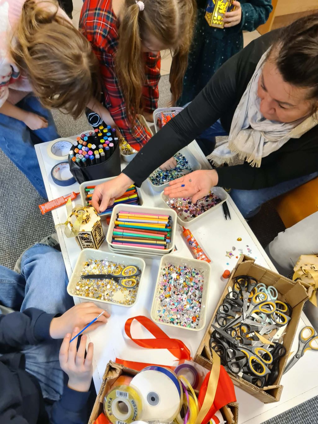 Dzieci i dorośli wspólnie tworzą świąteczne lampiony przy stołach warsztatowych.