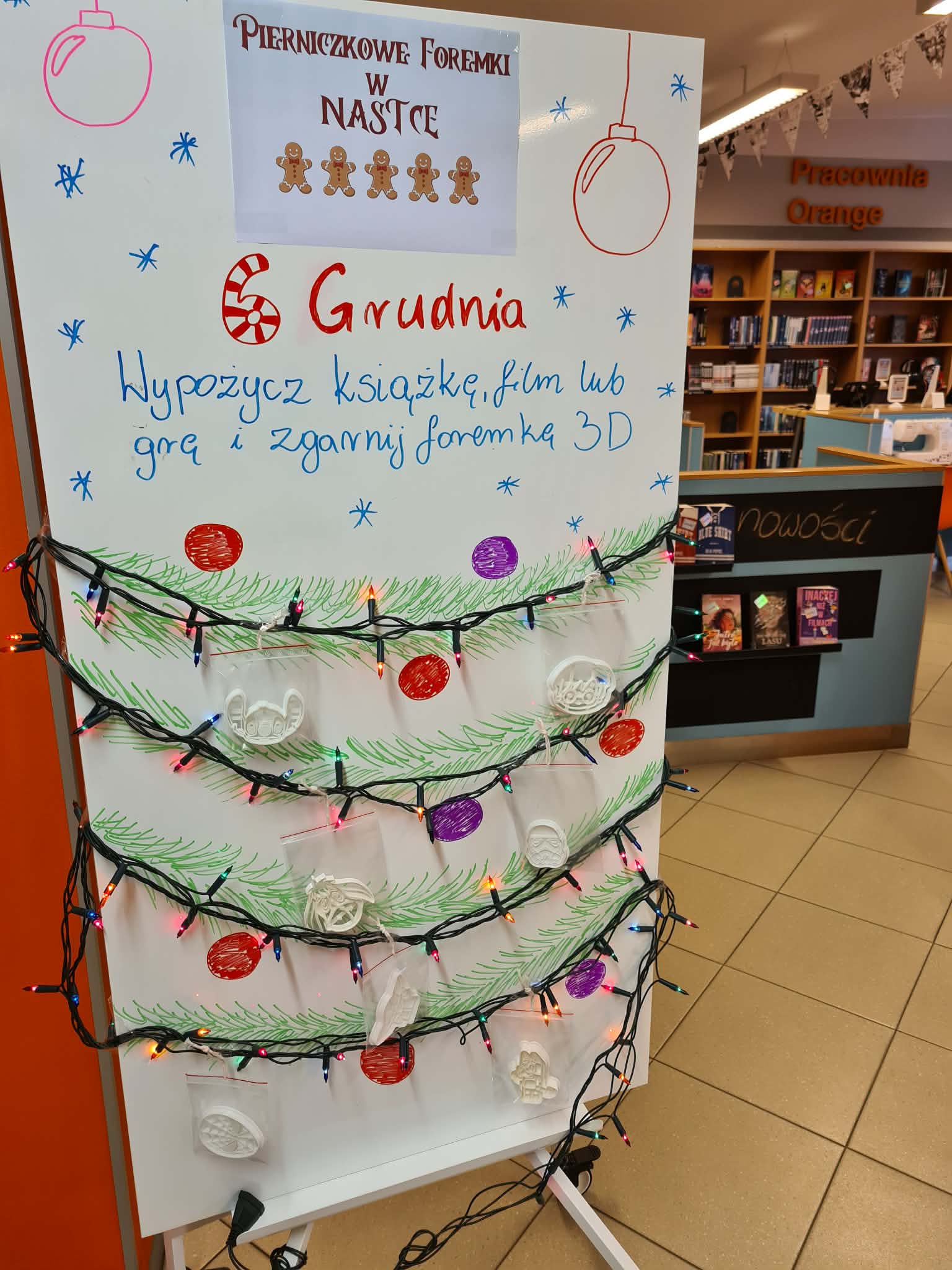 Tablica z napisem „Pierniczkowe foremki w Nastce”, udekorowana światełkami i rysunkową choinką. Na niej wiszą wydrukowane w 3D foremki do pierników.