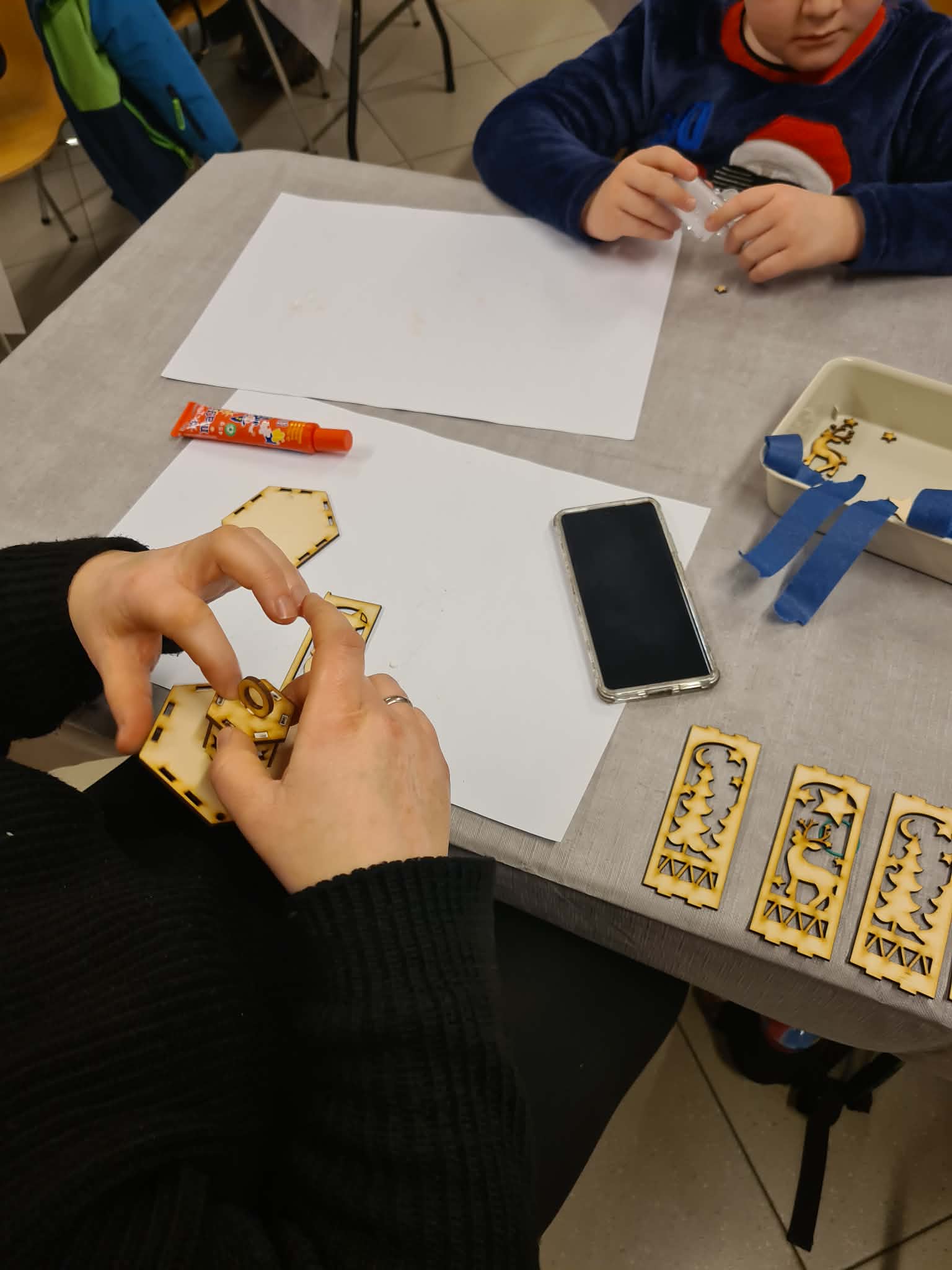 Dorośli i dzieci wspólnie montują lampion przy stole.