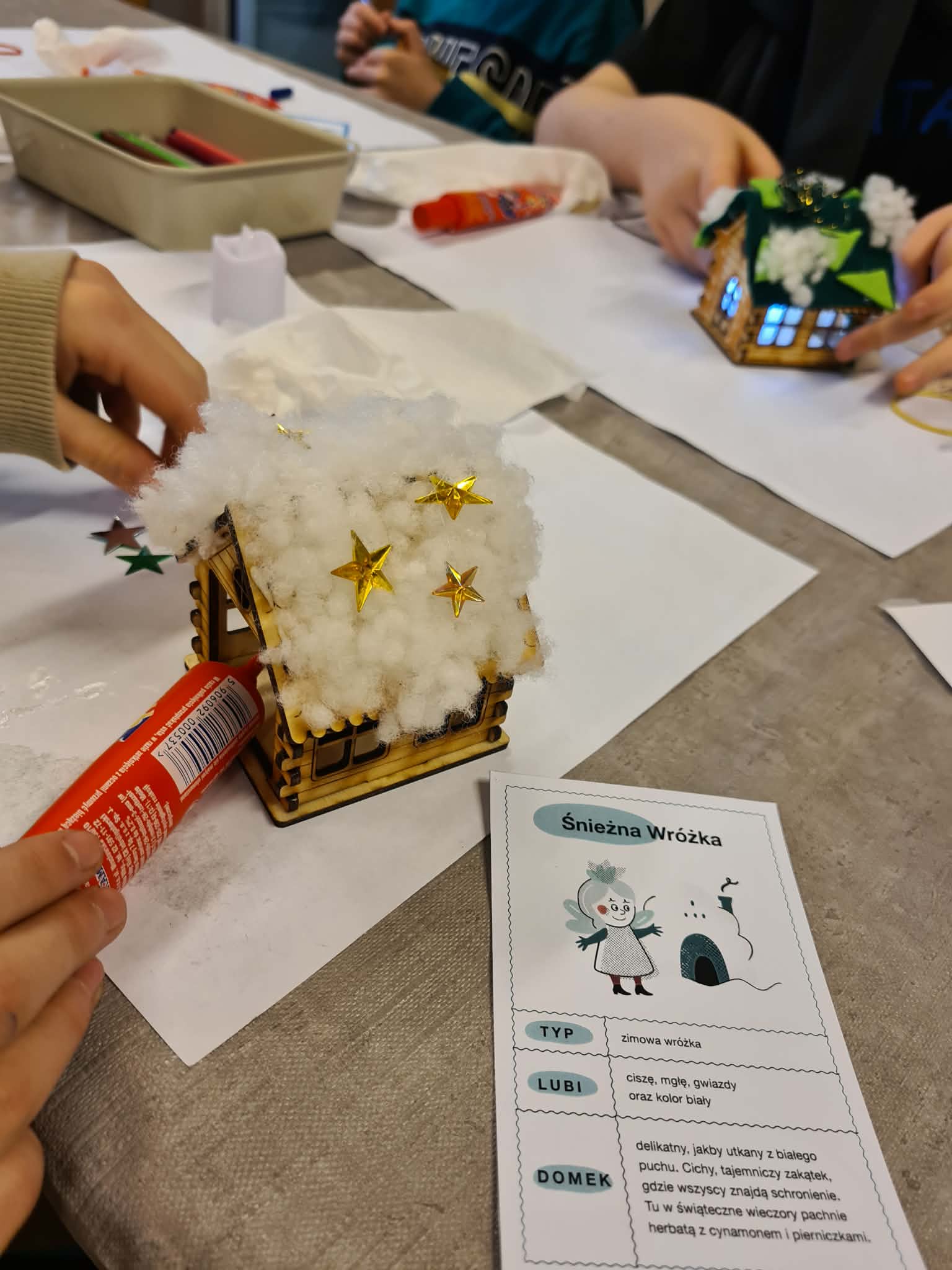 Dziecko nakleja złote gwiazdki na dach drewnianego domku pokrytego watą.
