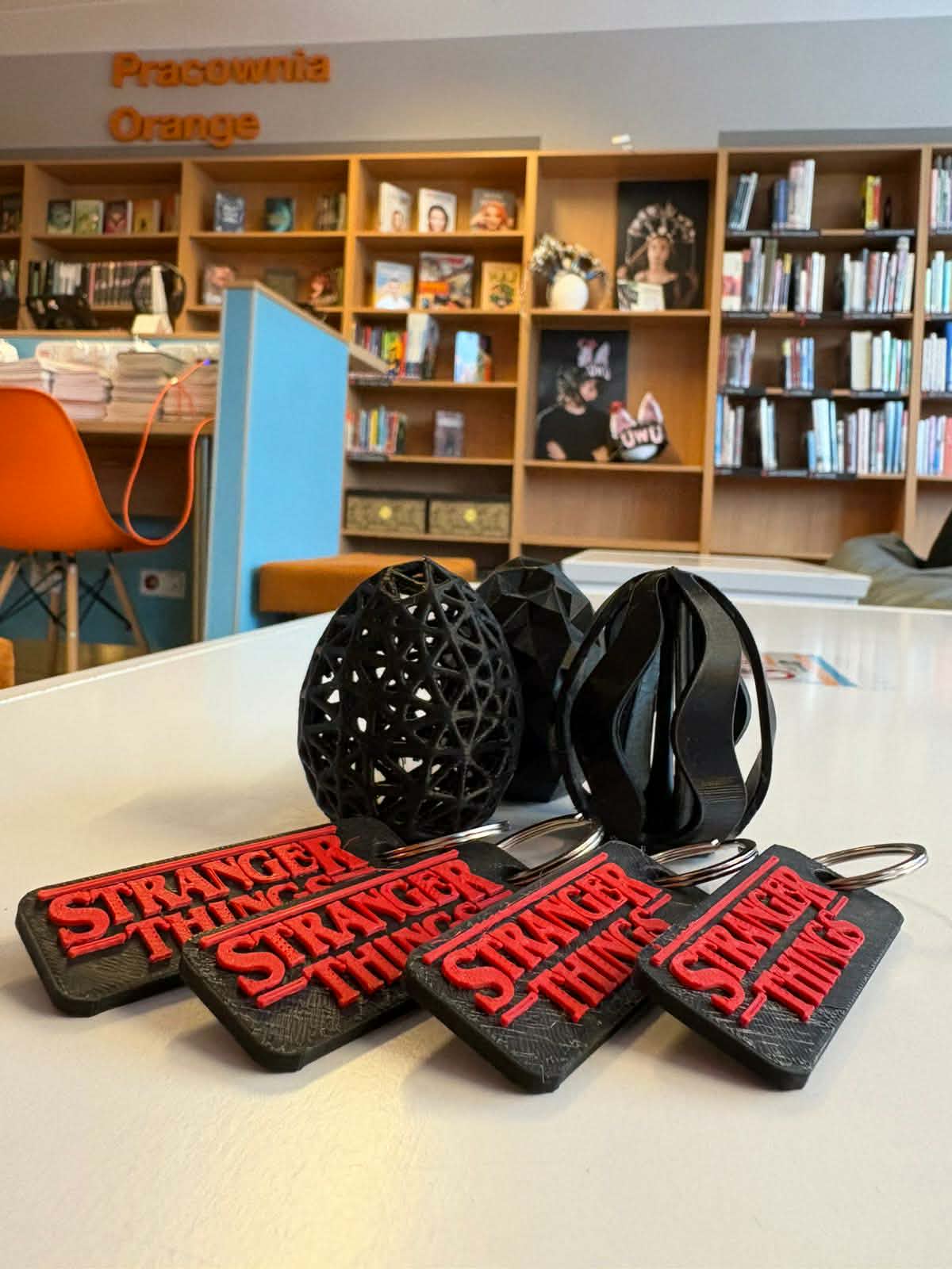 „Na stole leżą breloczki Stranger Things oraz czarne wydruki 3D; w tle widać regały i część Pracowni Orange.