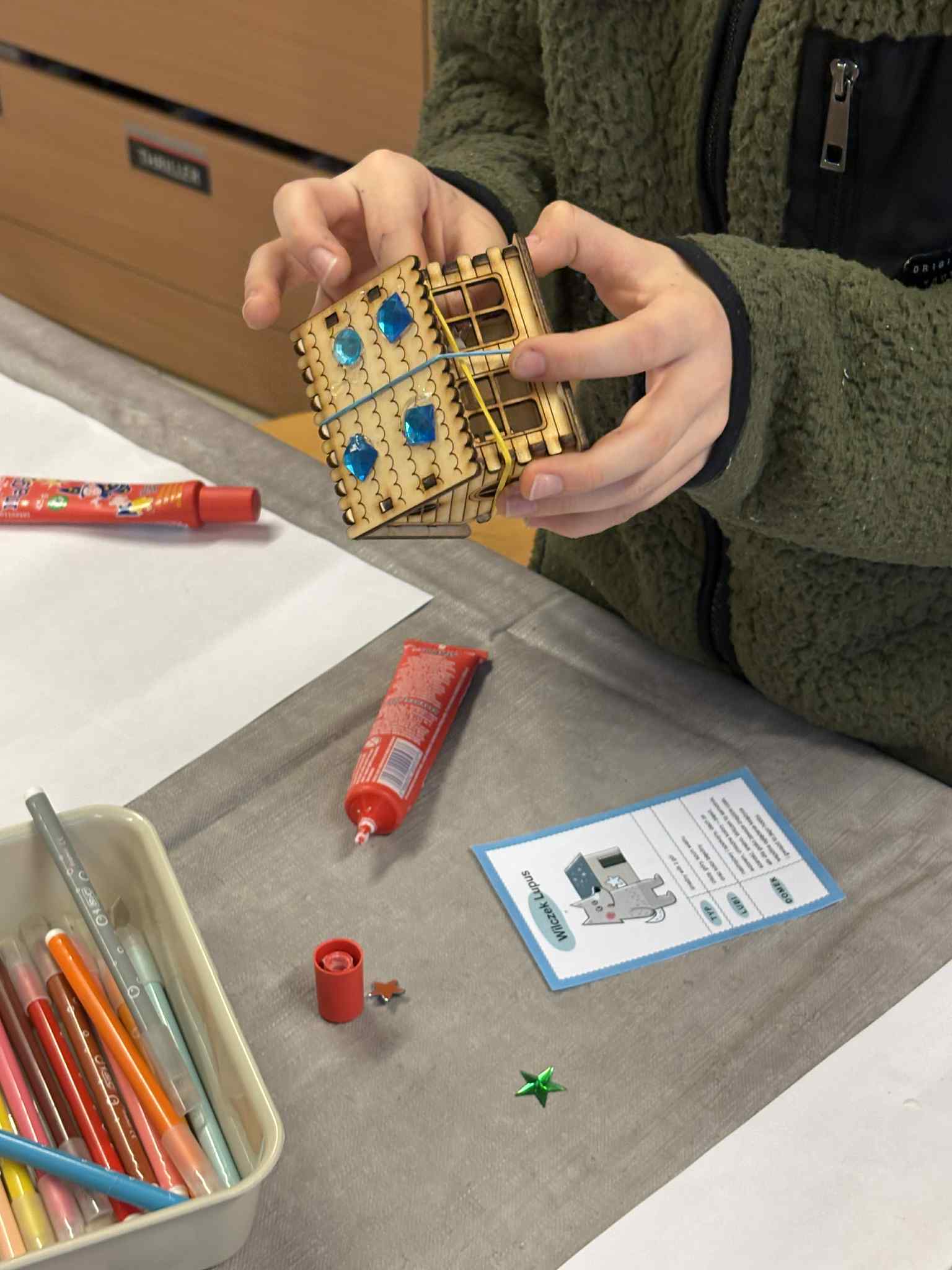 Dziecko składa drewniany domek, dociskając elementy, obok klej i niebieskie dekoracje.