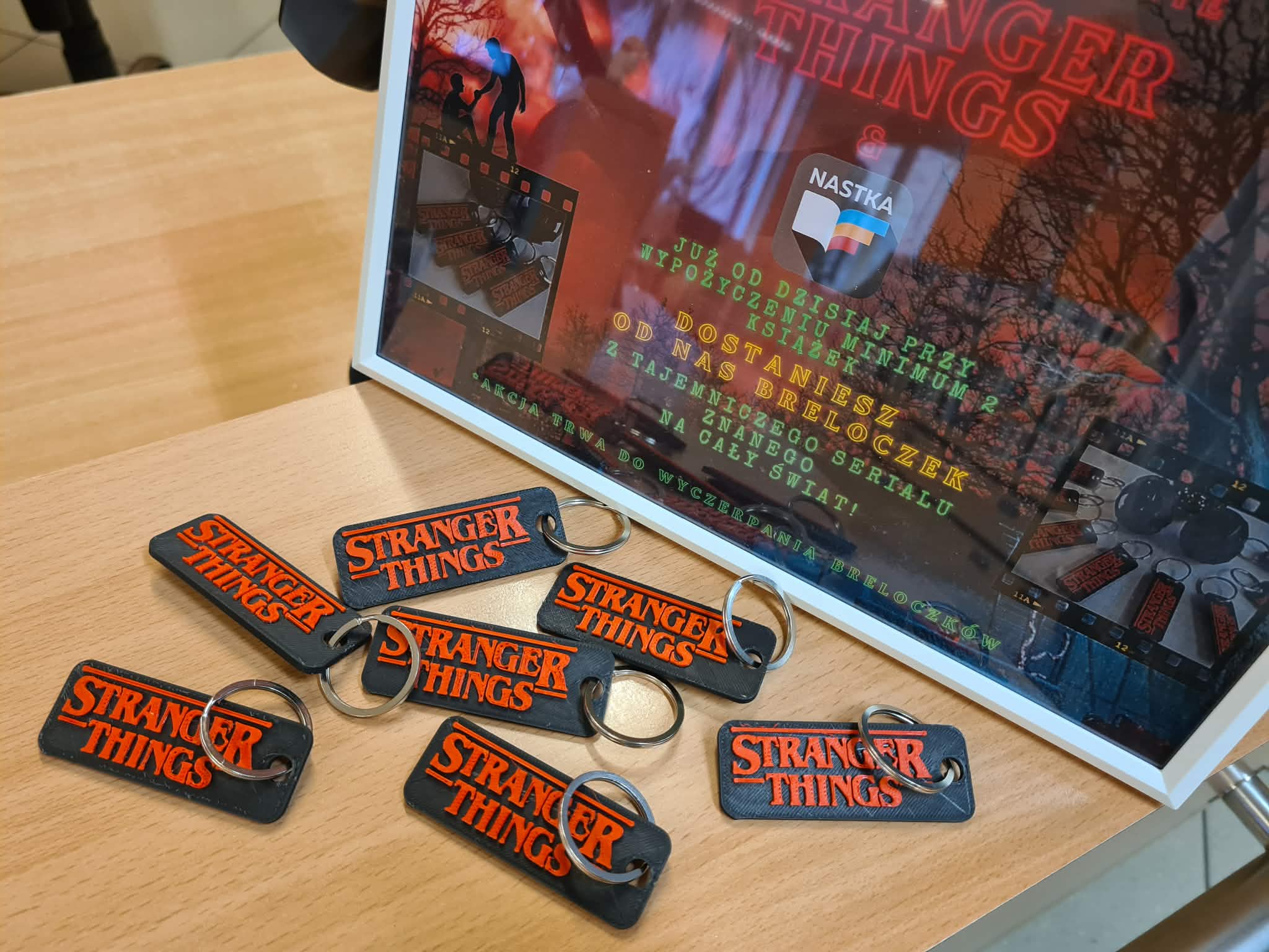 „Breloczki Stranger Things ułożone przed ramką z informacją o akcji promocyjnej w dziale młodzieżowym.