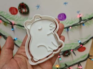 Foremka 3D w kształcie kapibary trzymana w dłoni, w tle tablica świąteczna ozdobiona lampkami.