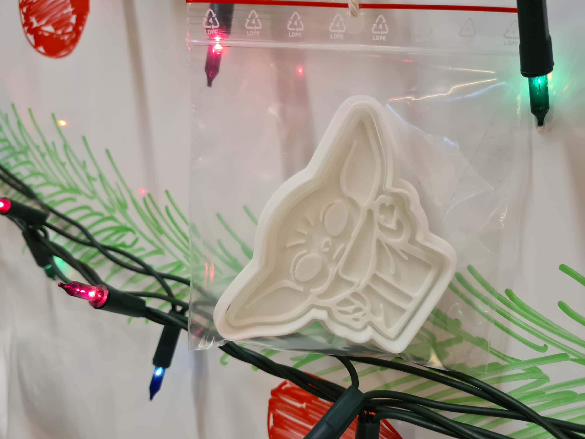 Foremka 3D w kształcie Baby Yody w foliowym opakowaniu, zawieszona na tablicy ze świątecznymi lampkami.