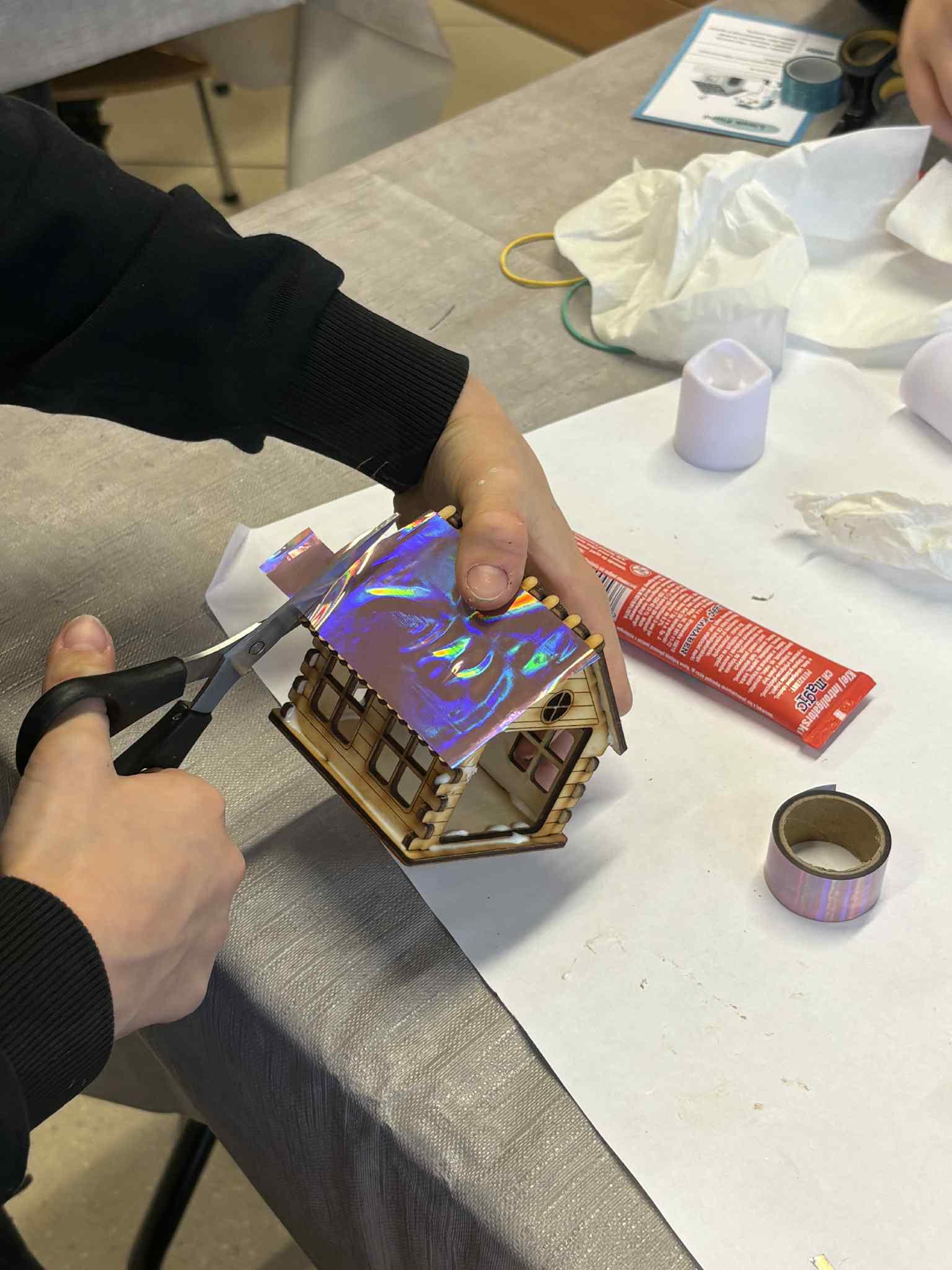 Uczestnik warsztatów docina błyszczącą holograficzną folię, którą nakleja na dach drewnianego domku.