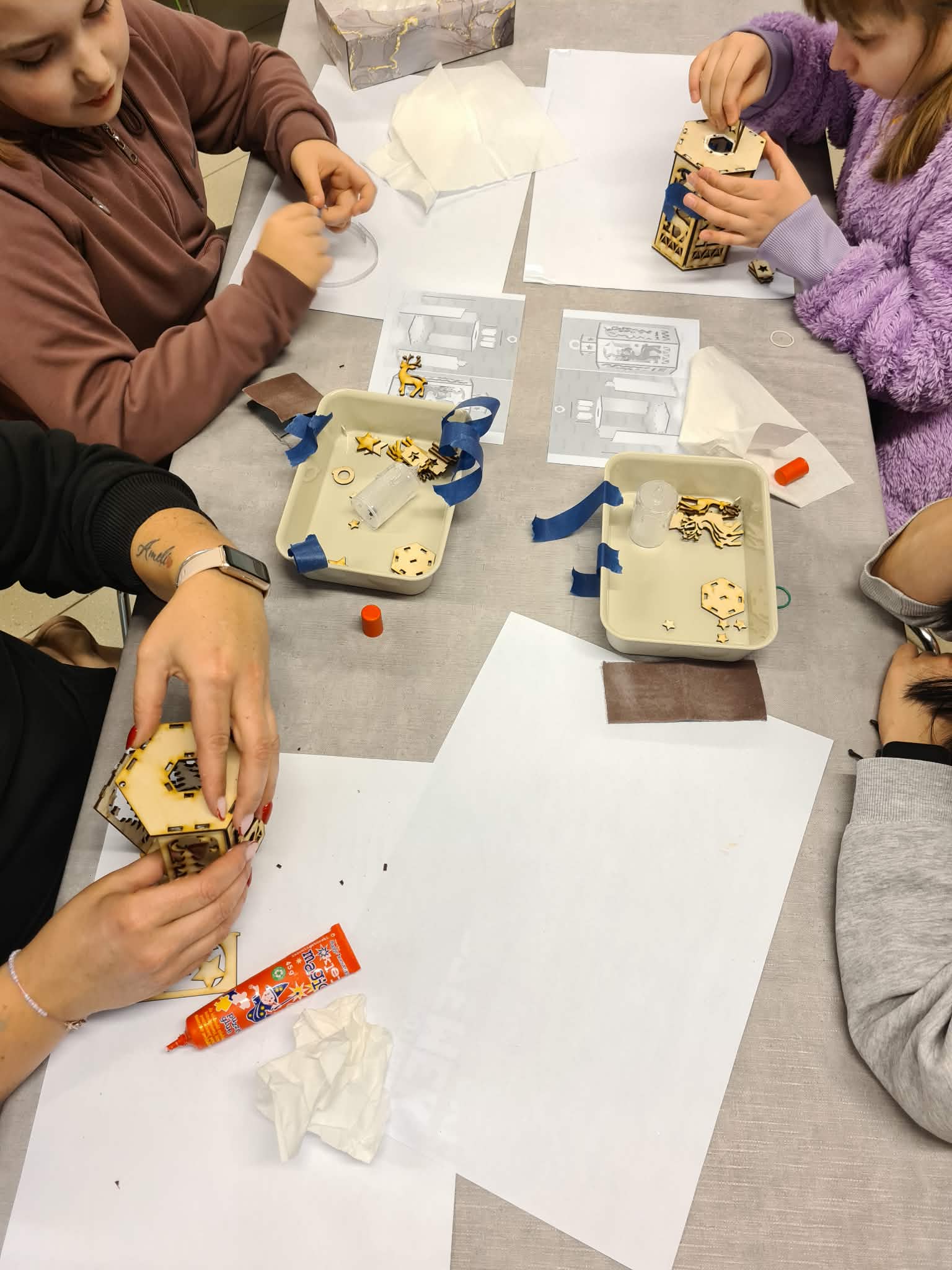 Dorośli i dzieci wspólnie montują lampion przy stole.