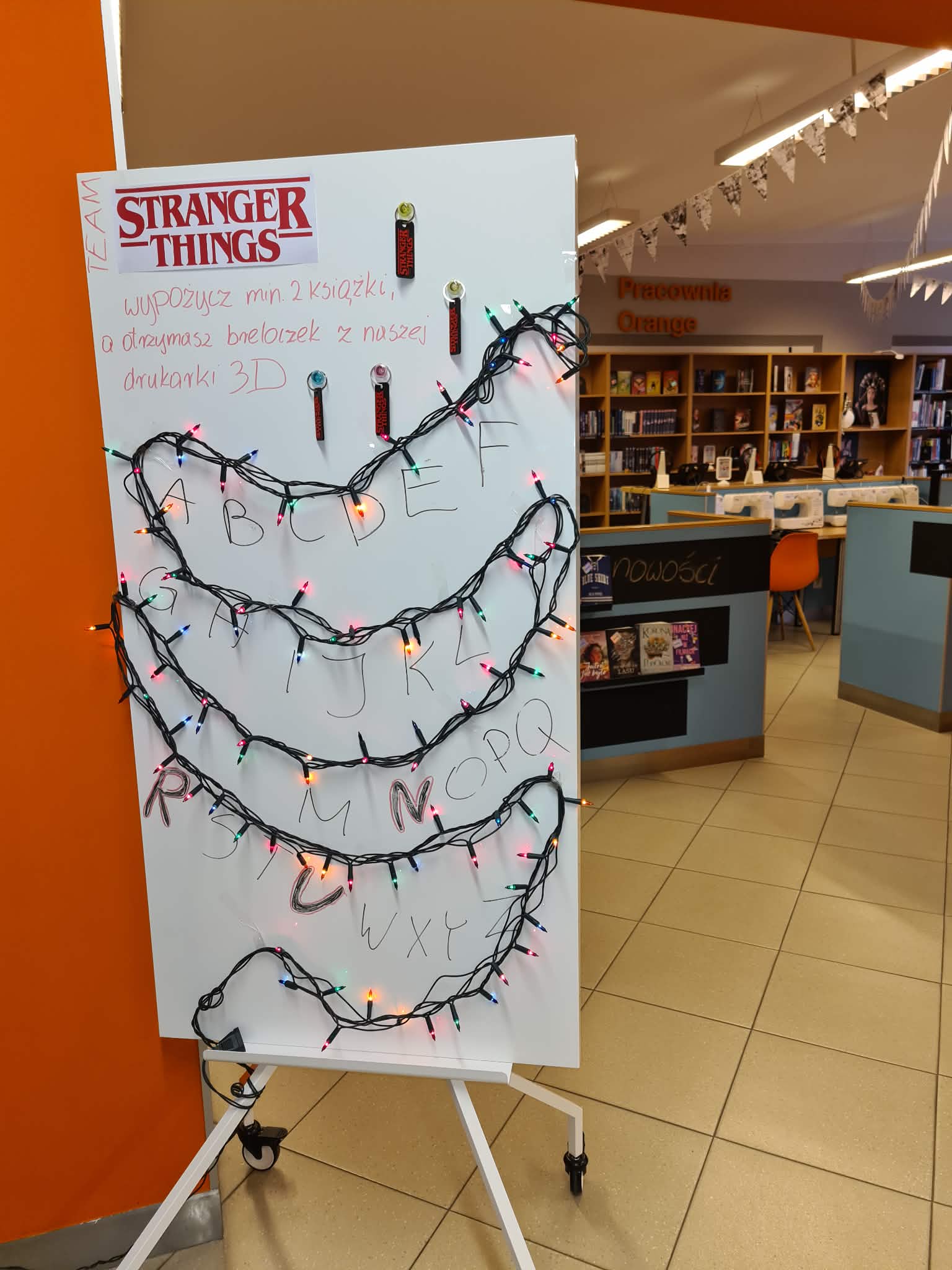 „Biała tablica ozdobiona kolorowymi lampkami i breloczkami Stranger Things, z ręcznie dopisanym tekstem o promocji.