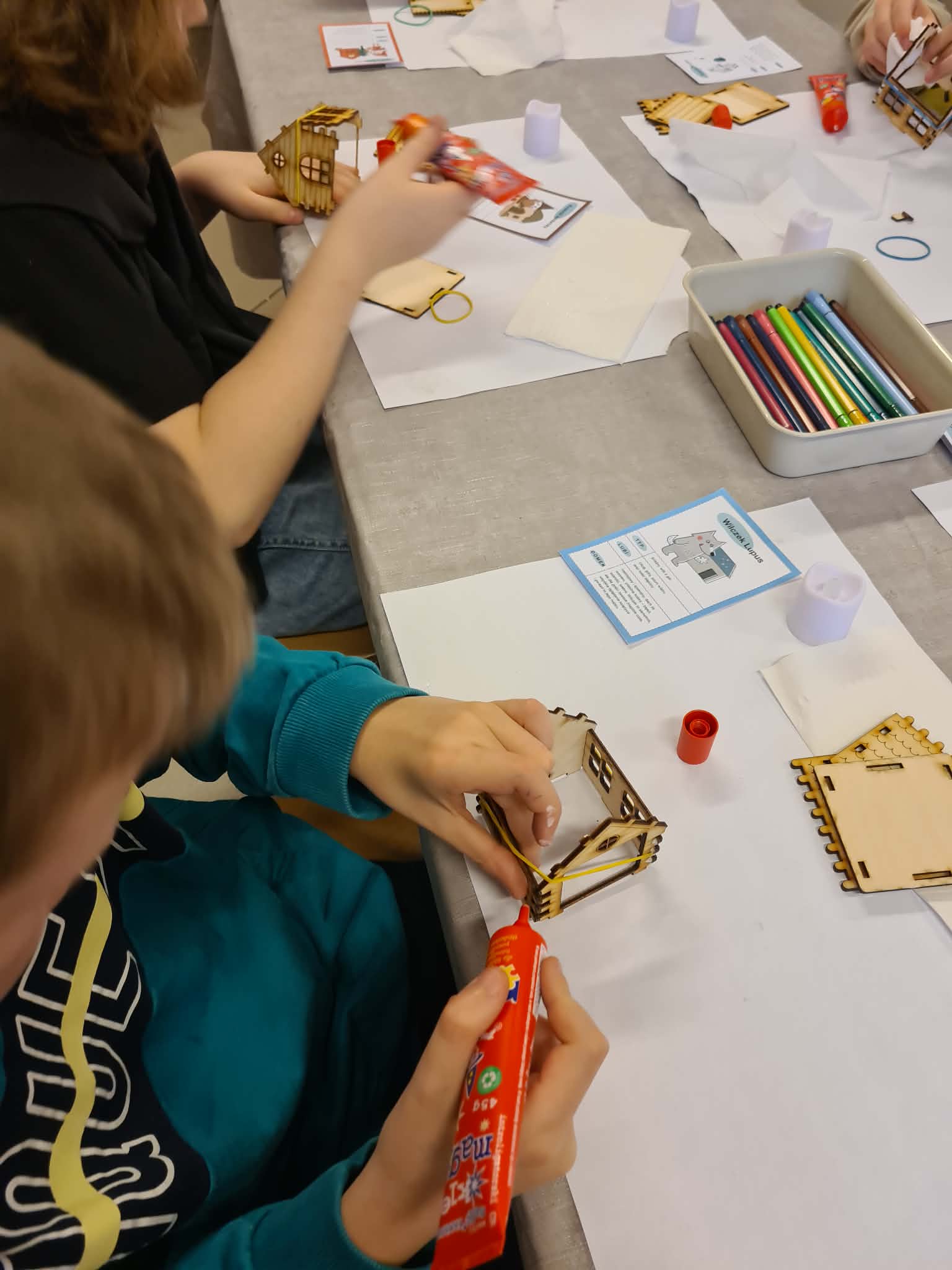 Dziecko klei elementy drewnianego domku, na stole leżą kredki i instrukcja z postacią wróżki.