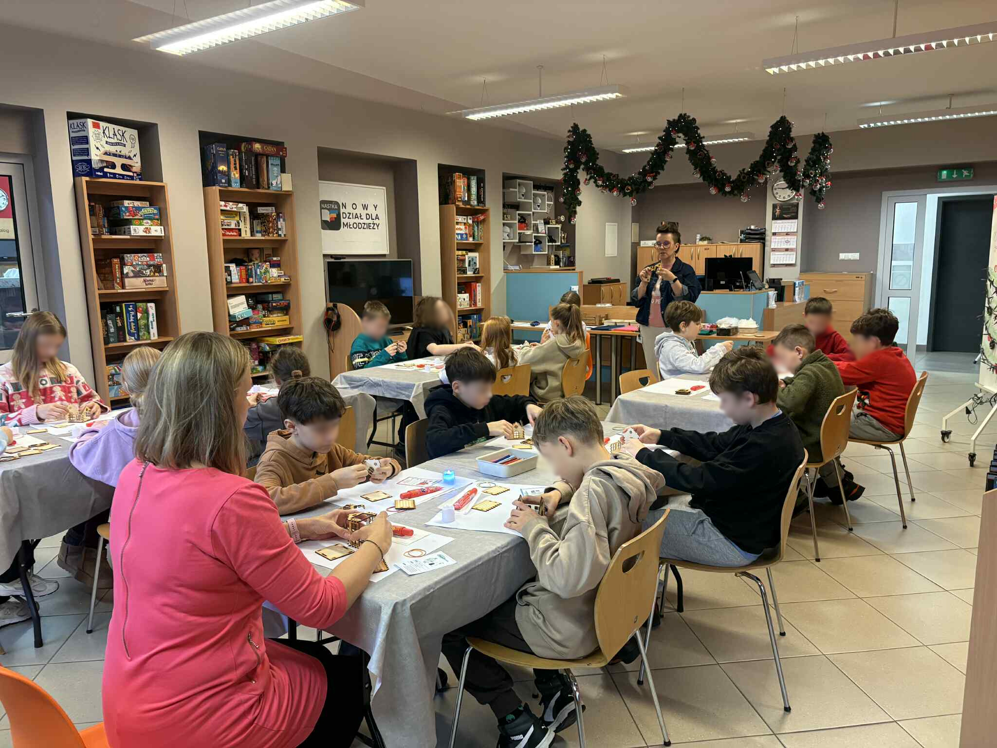 Grupa dzieci siedzi przy stołach w bibliotece i tworzy świąteczne domki pod okiem instruktorki.