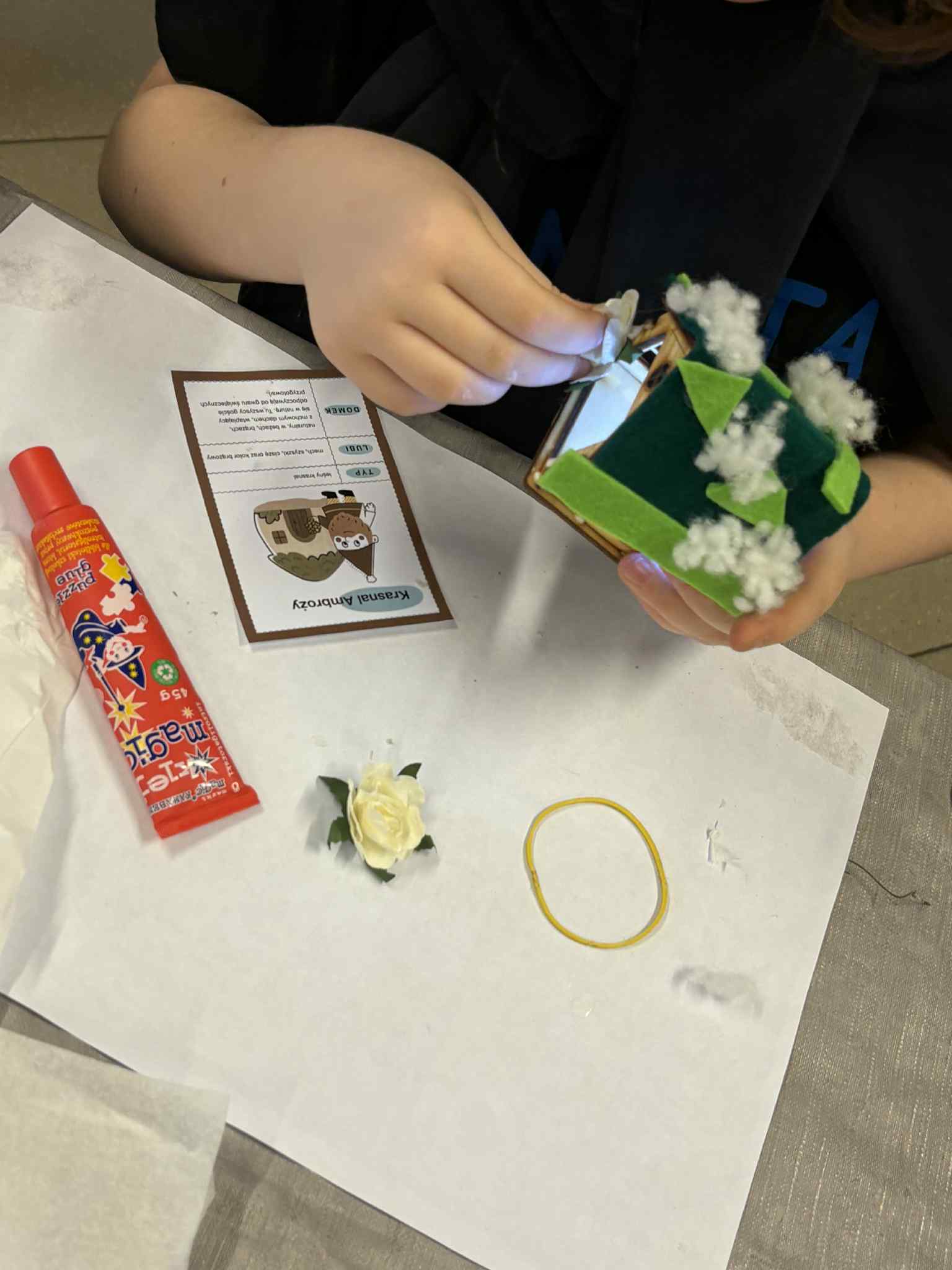 Dziecko przykleja biały kwiatek na dach zielonego domku z watą imitującą śnieg.