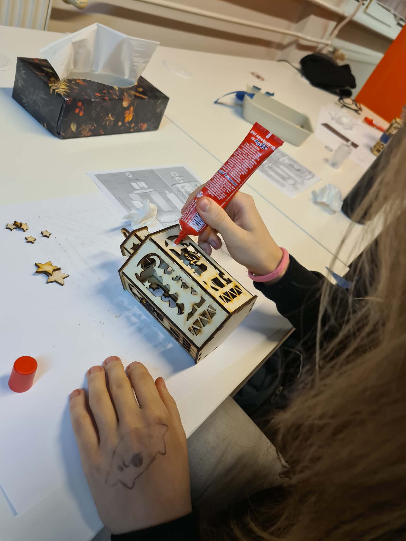 Dziecko wylewa klej z tubki na złożoną latarenkę, która leży na stole