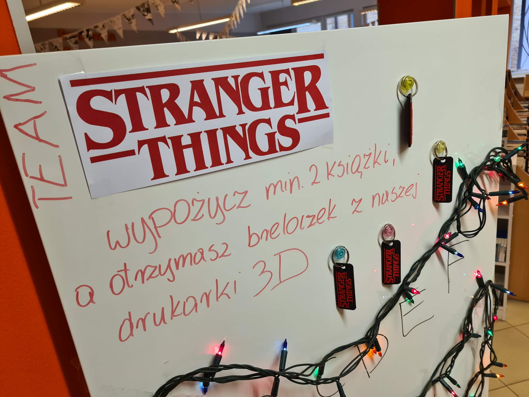 „Zbliżenie na tablicę z logo Stranger Things, kilkoma breloczkami oraz napisem o wypożyczeniu dwóch książek.