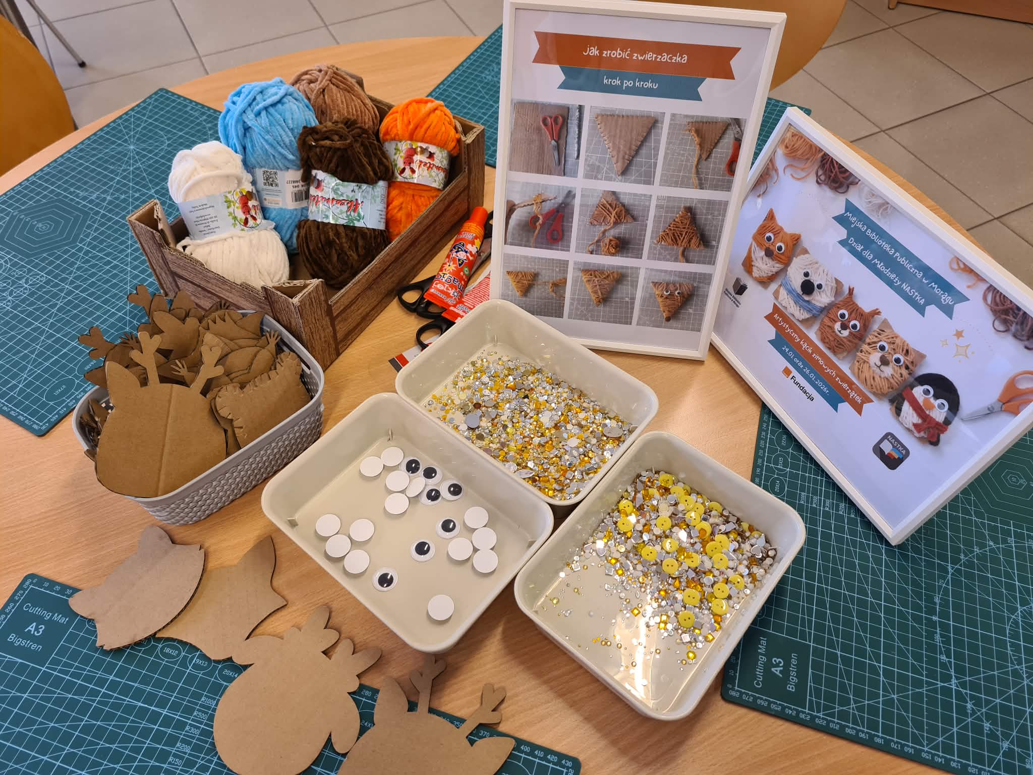 Materiały do warsztatów rękodzielniczych: włóczki, kartonowe kształty, oczy i ozdoby na stole.