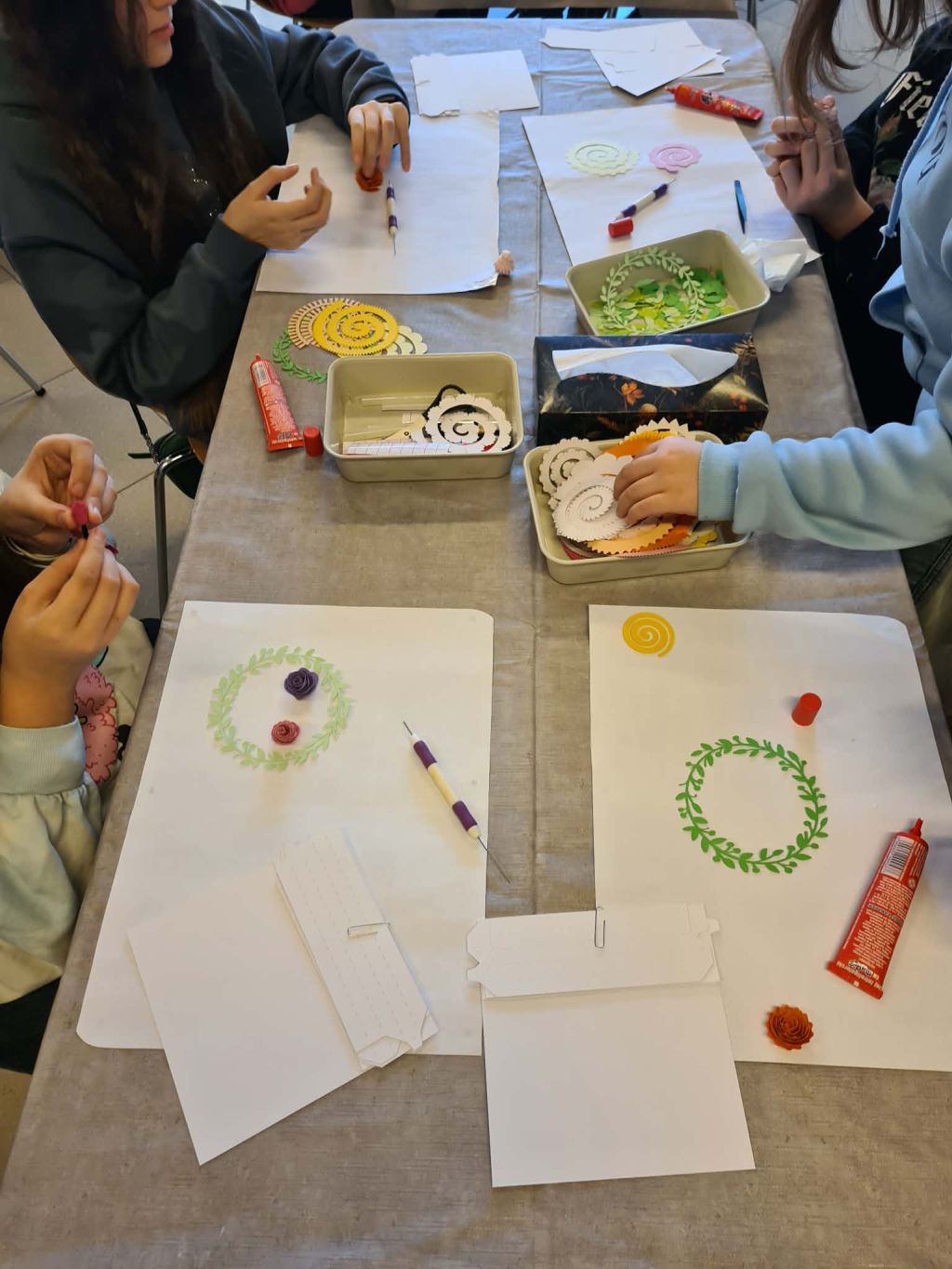 Dzieci przy stole tworzą papierowe kwiaty, na blacie narzędzia i elementy dekoracyjne.