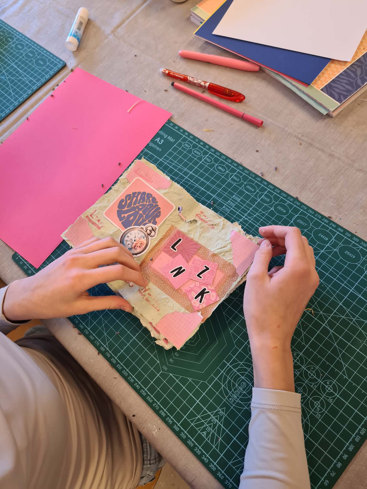 Dłonie układają ozdobne litery i elementy na okładce książeczki z papieru czerpanego.