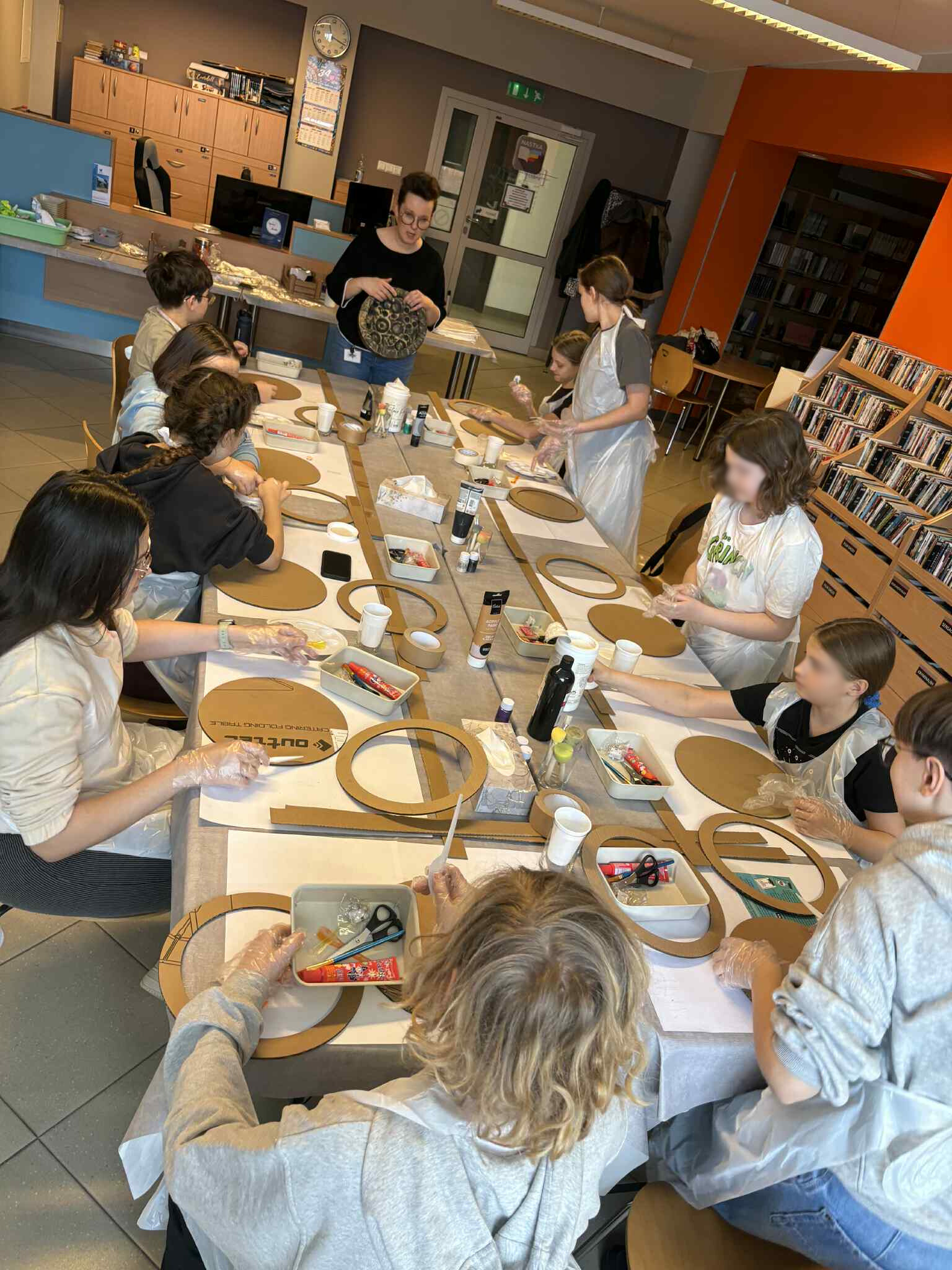 Prowadząca pokazuje pracę plastyczną grupie przy stole.