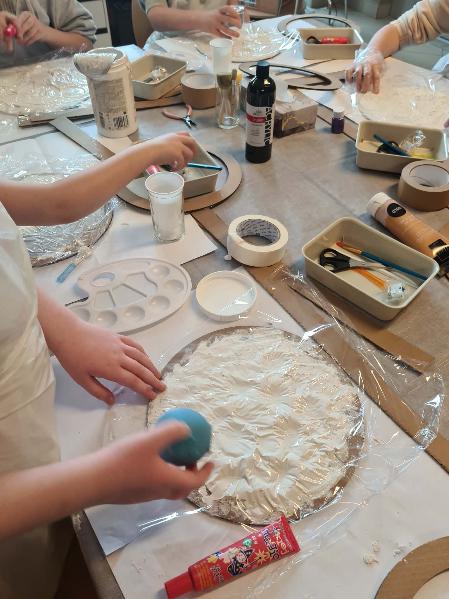 Dzieci przy stole warsztatowym, przygotowują okrągłą formę pokrytą masą plastyczną.