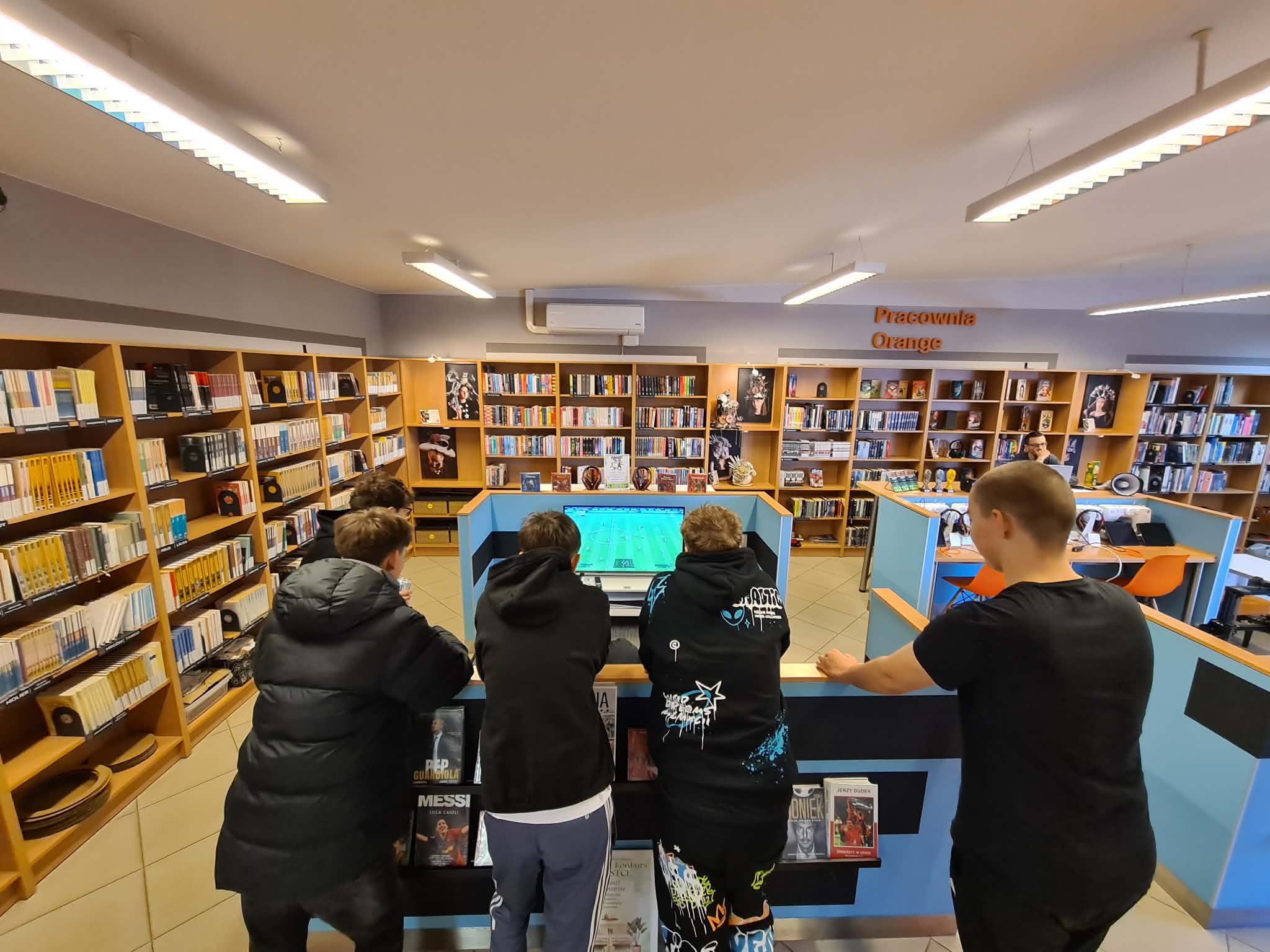 Grupa młodzieży opiera się o ladę w bibliotece, wspólnie oglądając mecz piłkarski wyświetlany na ekranie. W tle regały z książkami.