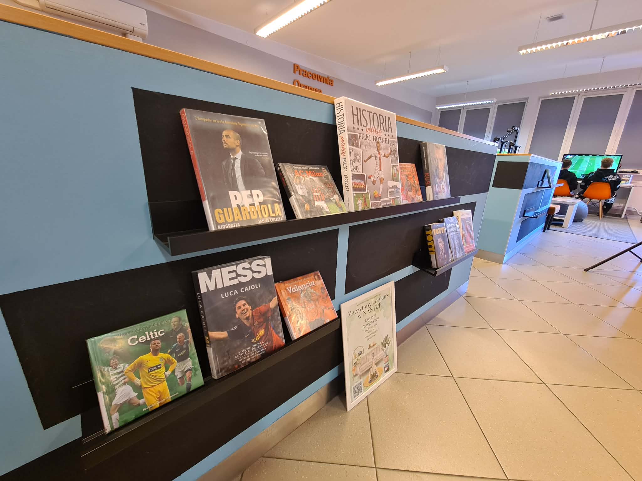 Ekspozycja książek o tematyce piłkarskiej i sportowej na ladzie w bibliotece, w tym biografie znanych piłkarzy. W tle widoczna sala z konsolami.