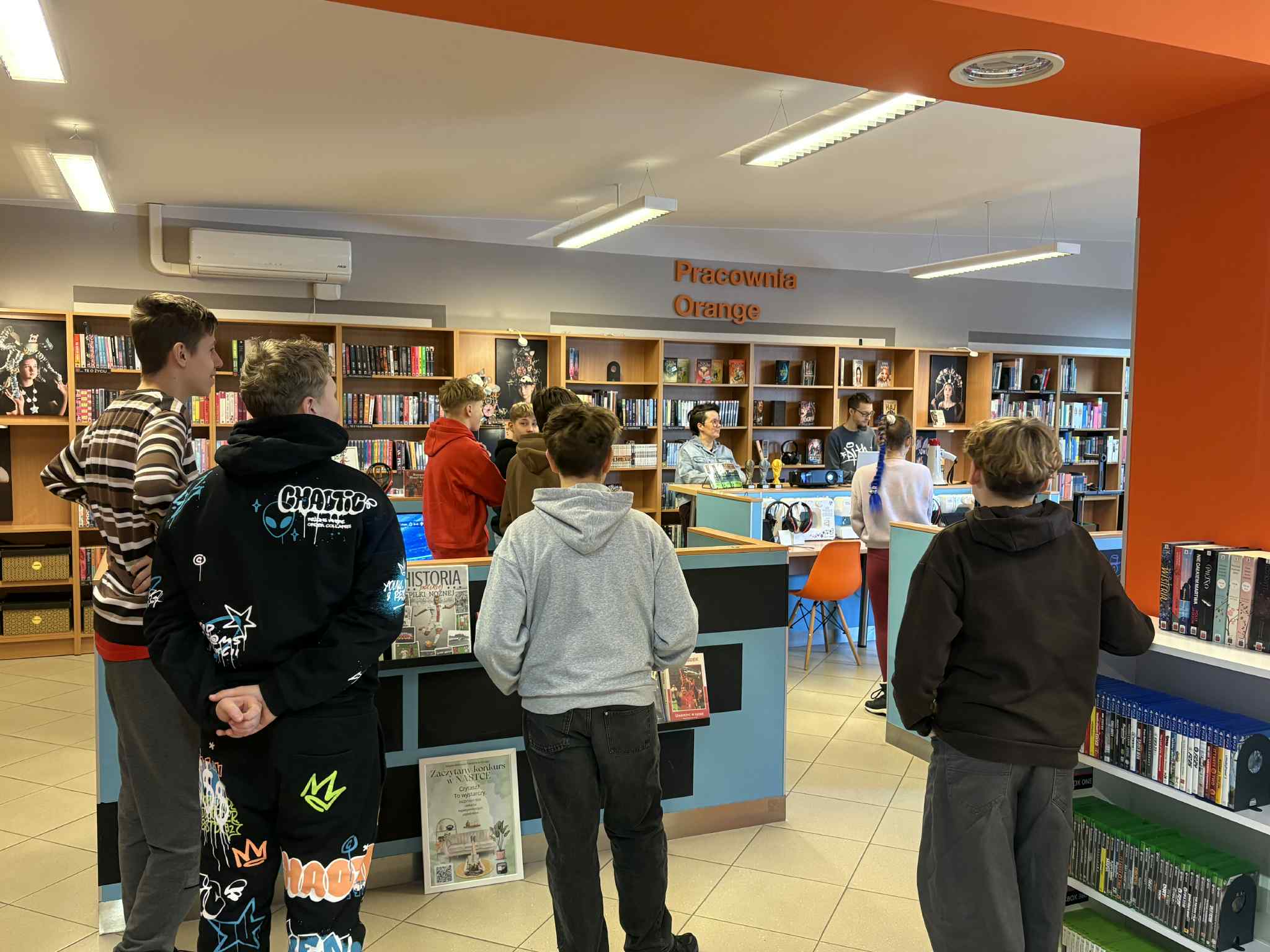 Grupa nastolatków stoi w Dziale dla Młodzieży NASTKA w bibliotece, oglądając mecz piłkarski na ekranie. W tle regały z książkami i napis „Pracownia Orange”