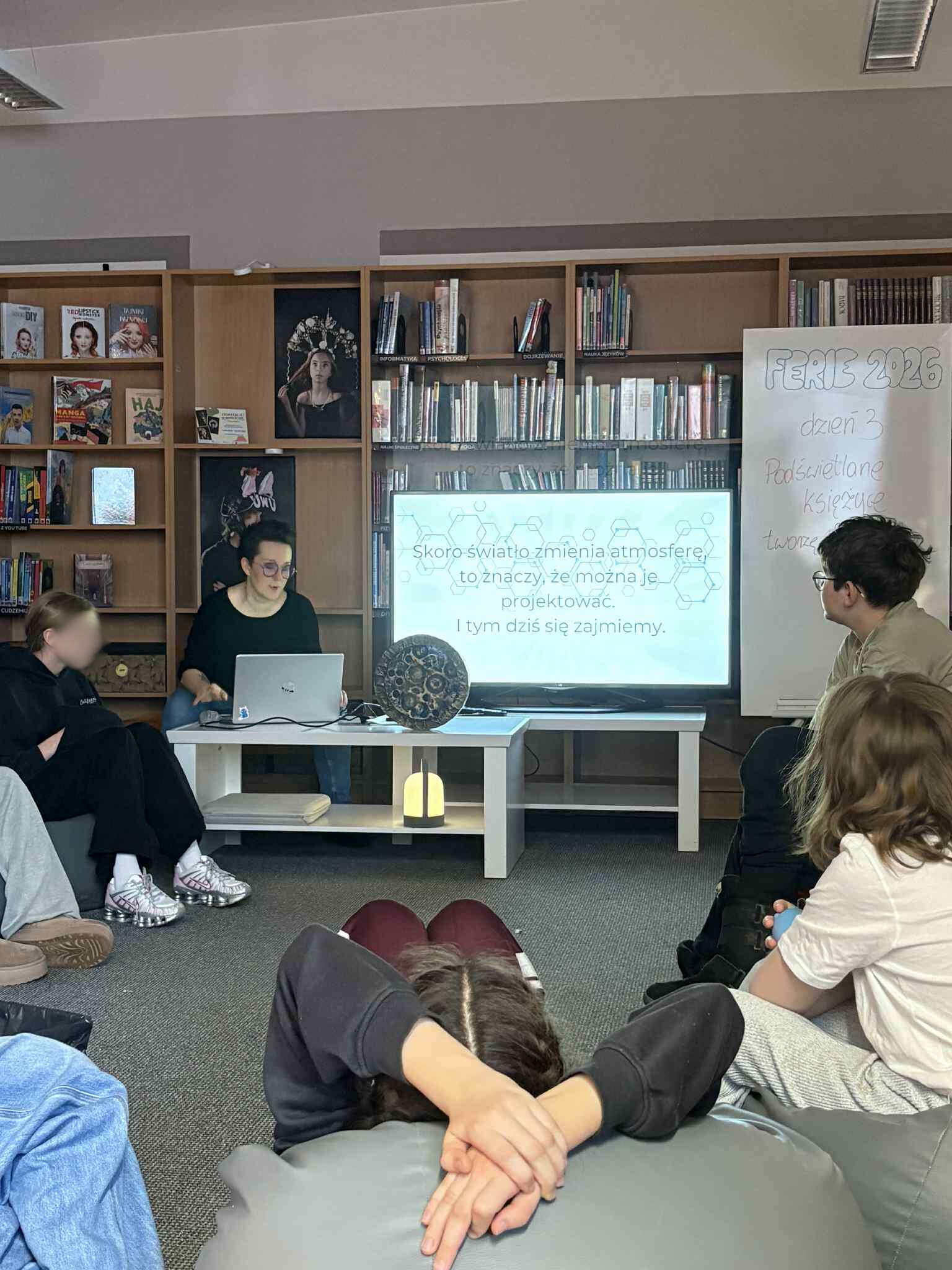Widok sali: grupa dzieci siedzi na pufach, w tle prowadząca i laptop.