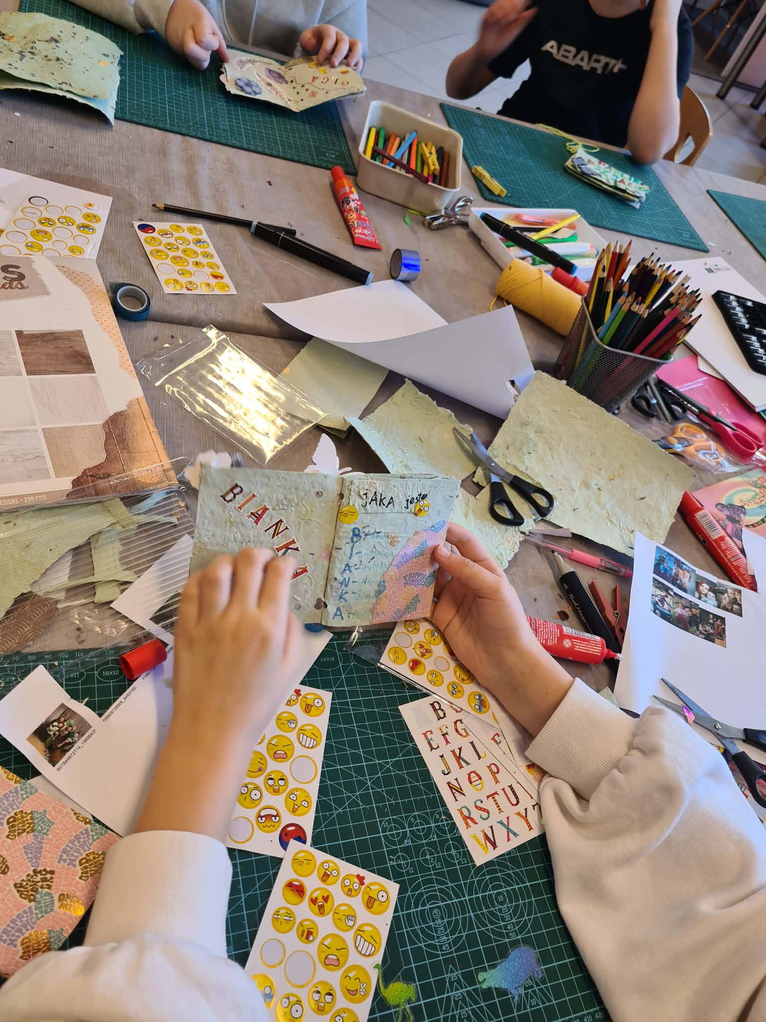 Ręce dzieci ozdabiają małe książeczki z papieru czerpanego przy stole pełnym materiałów plastycznych.