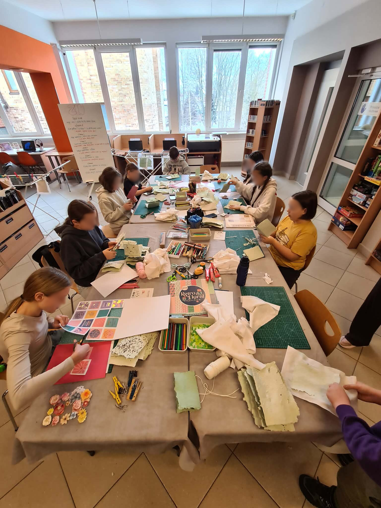 Uczestnicy warsztatów siedzą przy stole i pracują nad książeczkami z papieru czerpanego.