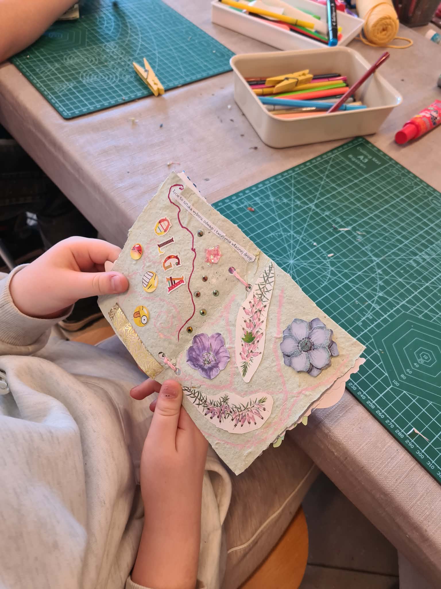 Dziecko prezentuje ozdobioną stronę książeczki z rysunkami, naklejkami i napisami.