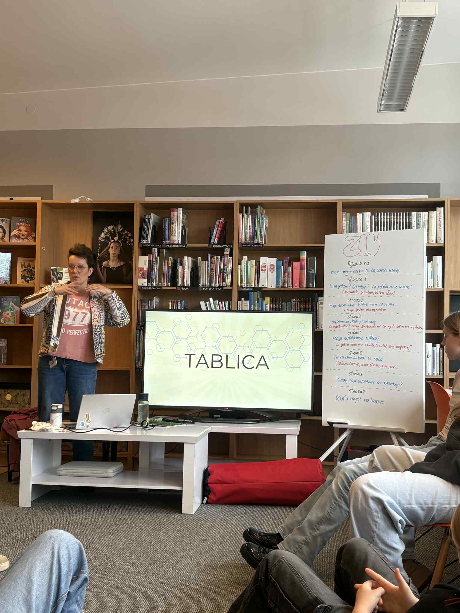 Prowadząca stoi przy ekranie i tablicy z zapisanymi punktami zajęć w bibliotece.