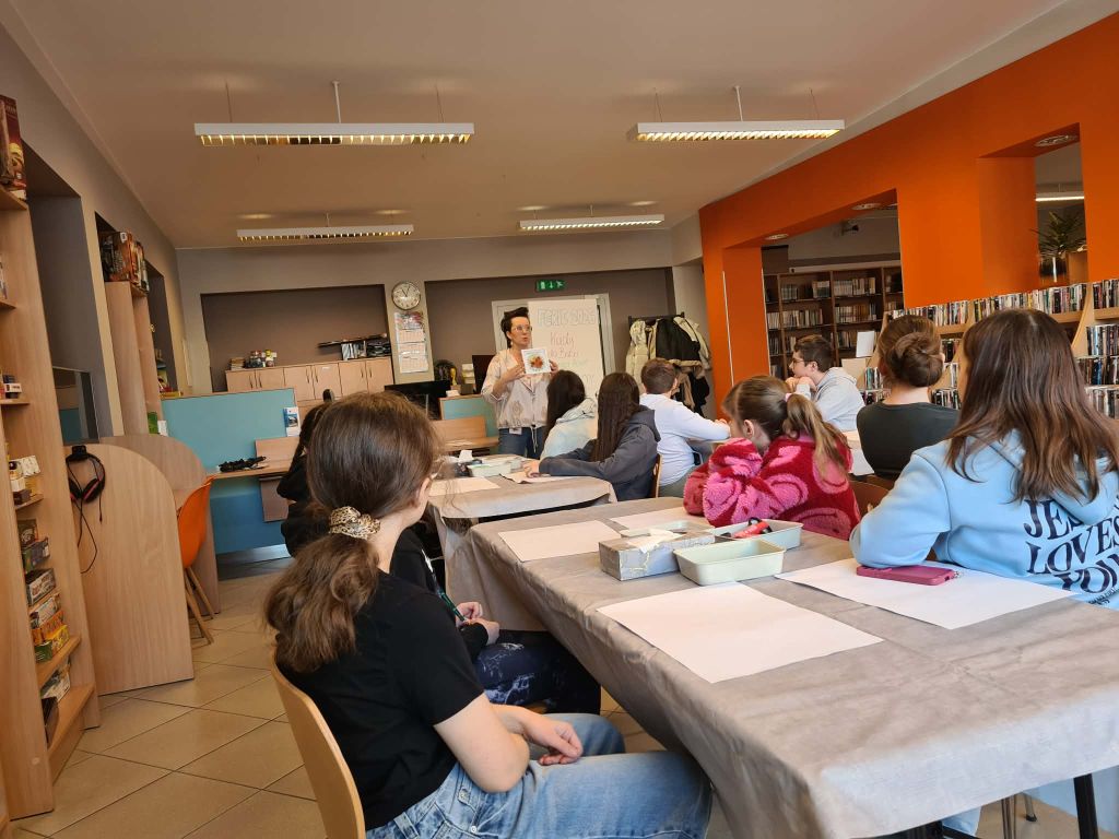 Uczestnicy warsztatów siedzą przy stołach w bibliotece, prowadząca prezentuje gotową pracę plastyczną.