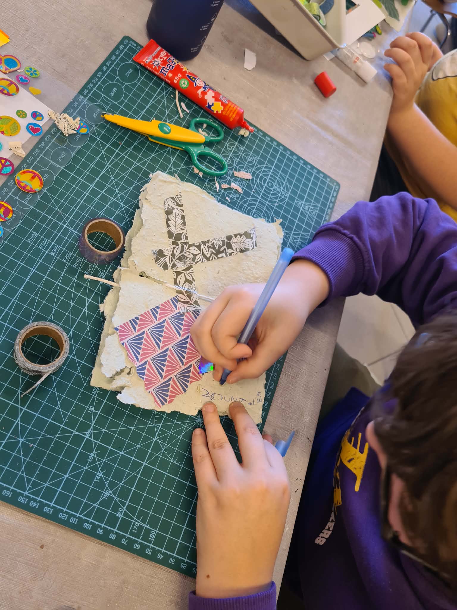 Dziecko pisze długopisem na kartce z papieru czerpanego, leżącej na macie do cięcia.