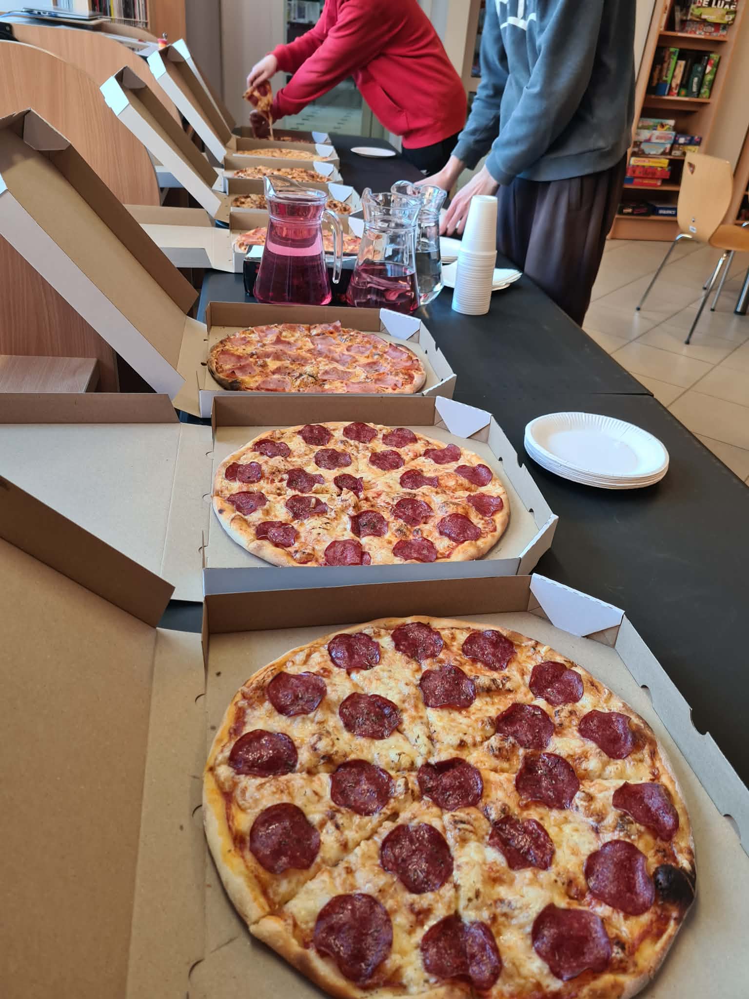 Stoły z otwartymi pudełkami pizzy, talerzykami i dzbankami z napojami przygotowane dla uczestników wydarzenia w bibliotece.