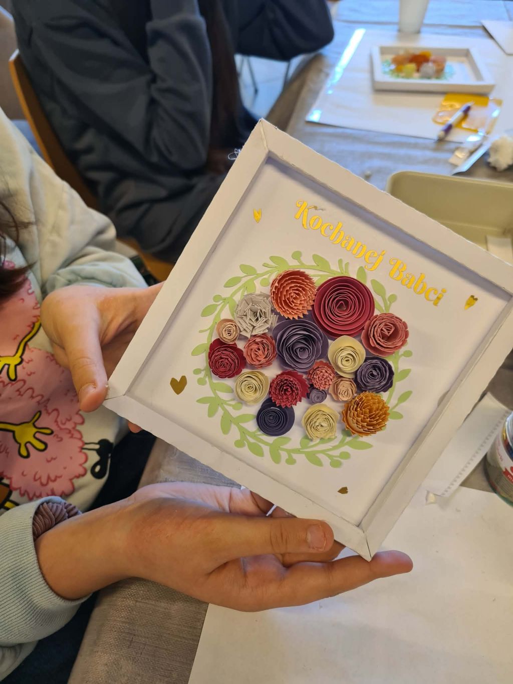 Zbliżenie na ramkę z papierowymi kwiatami i napisem „Kochanej Babci”, trzymaną w dłoniach dziecka.