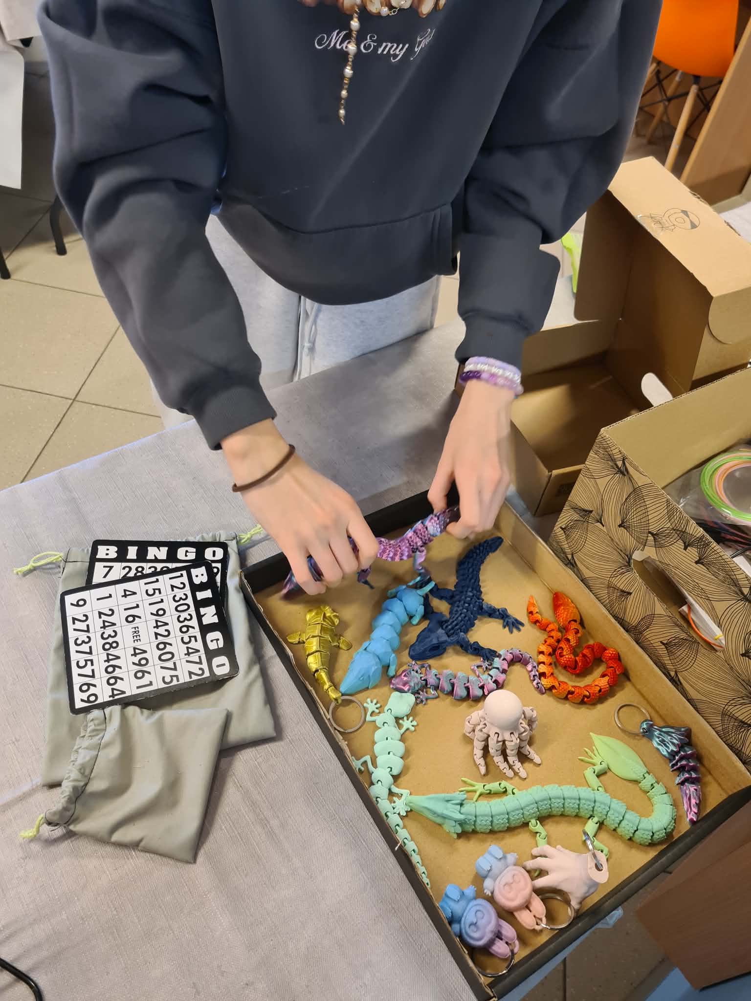 Dłonie osoby wybierają kolorowe figurki i breloki z pudełka na stole warsztatowym.