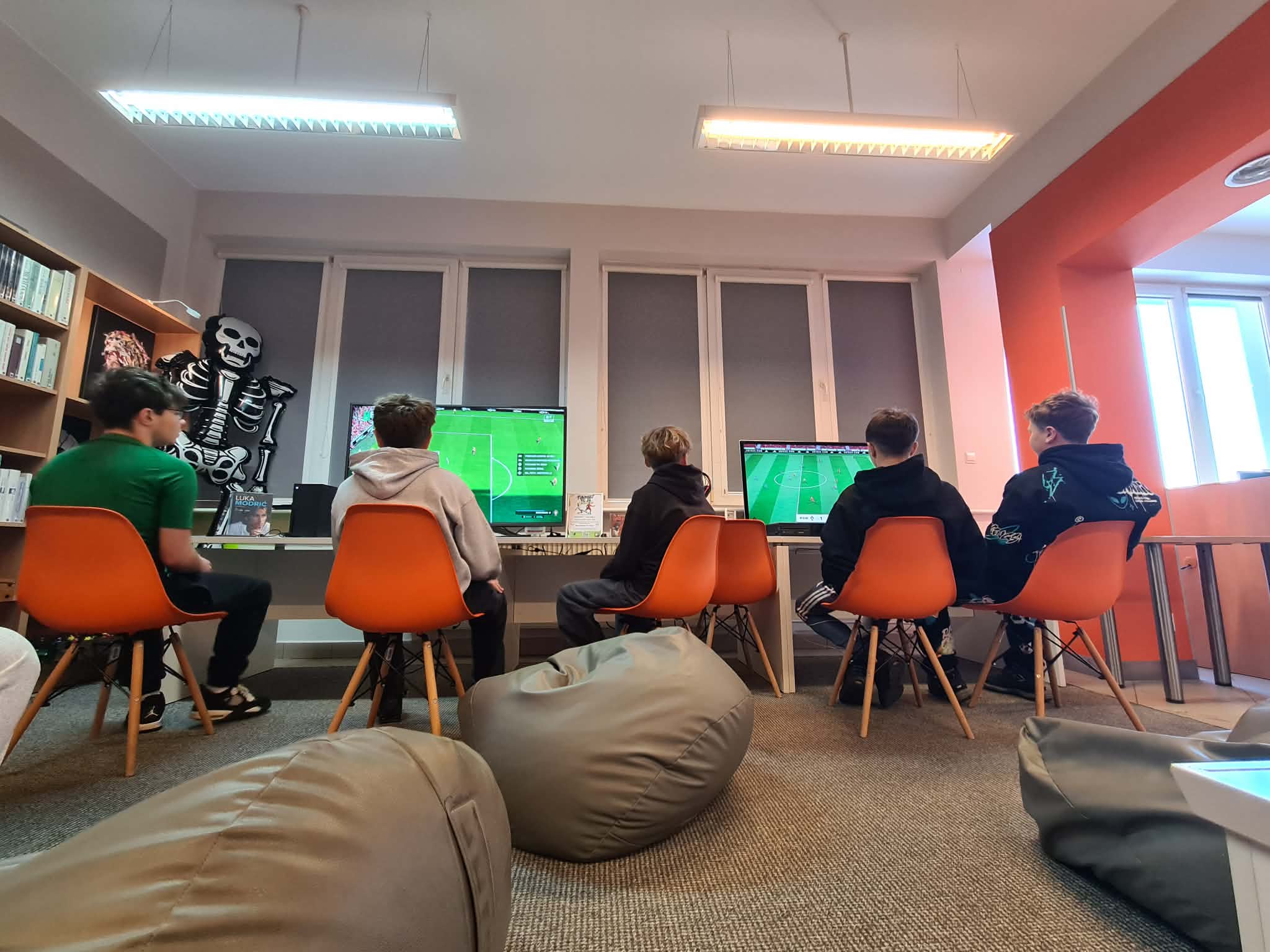 Kilku chłopców siedzi na pomarańczowych krzesłach przed monitorami i gra w grę piłkarską na konsolach. Na podłodze leżą pufy, w tle biblioteczne regały.