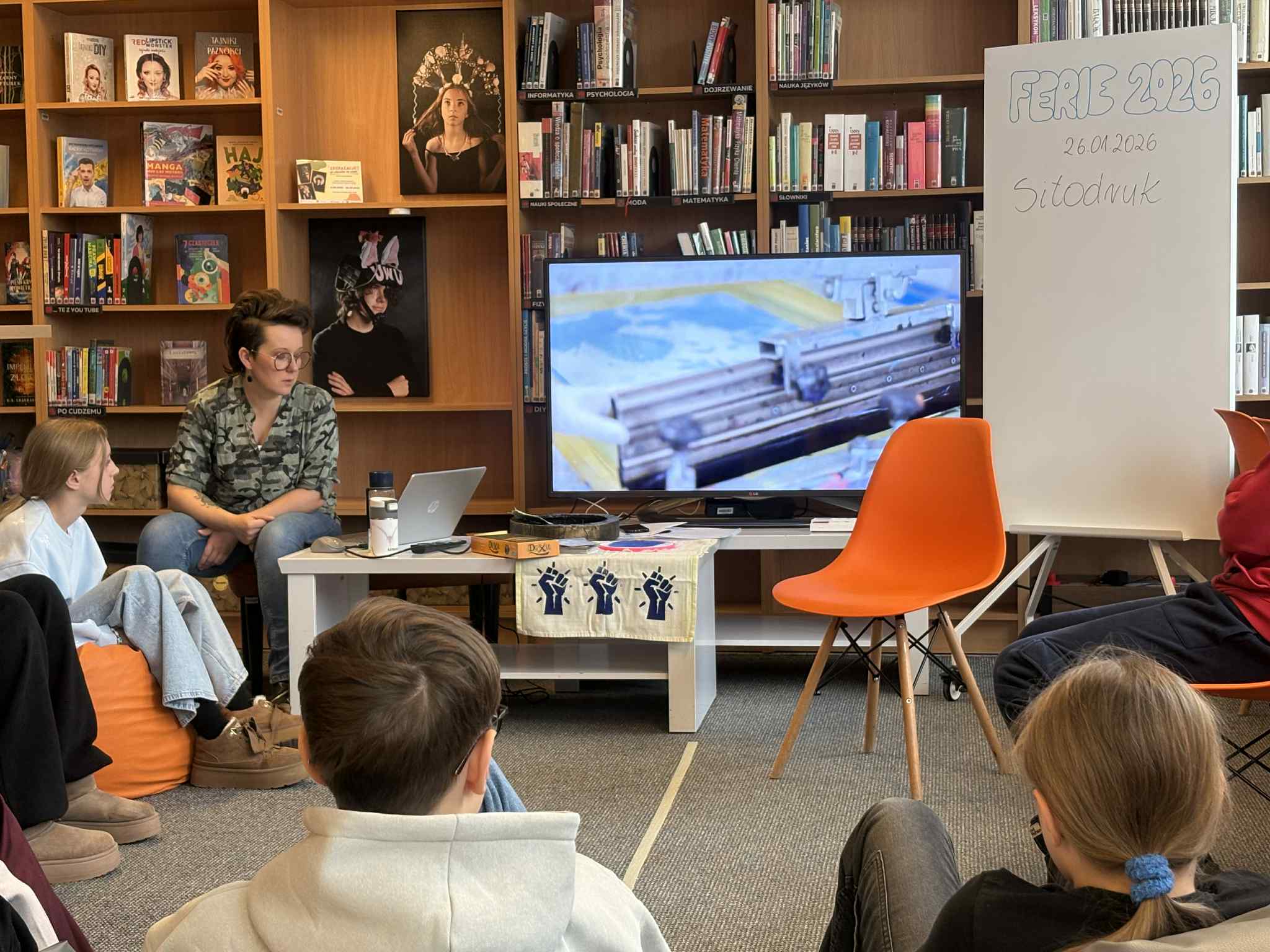 Uczestnicy warsztatów siedzą w bibliotece, prowadząca omawia zajęcia przy ekranie i tablicy.