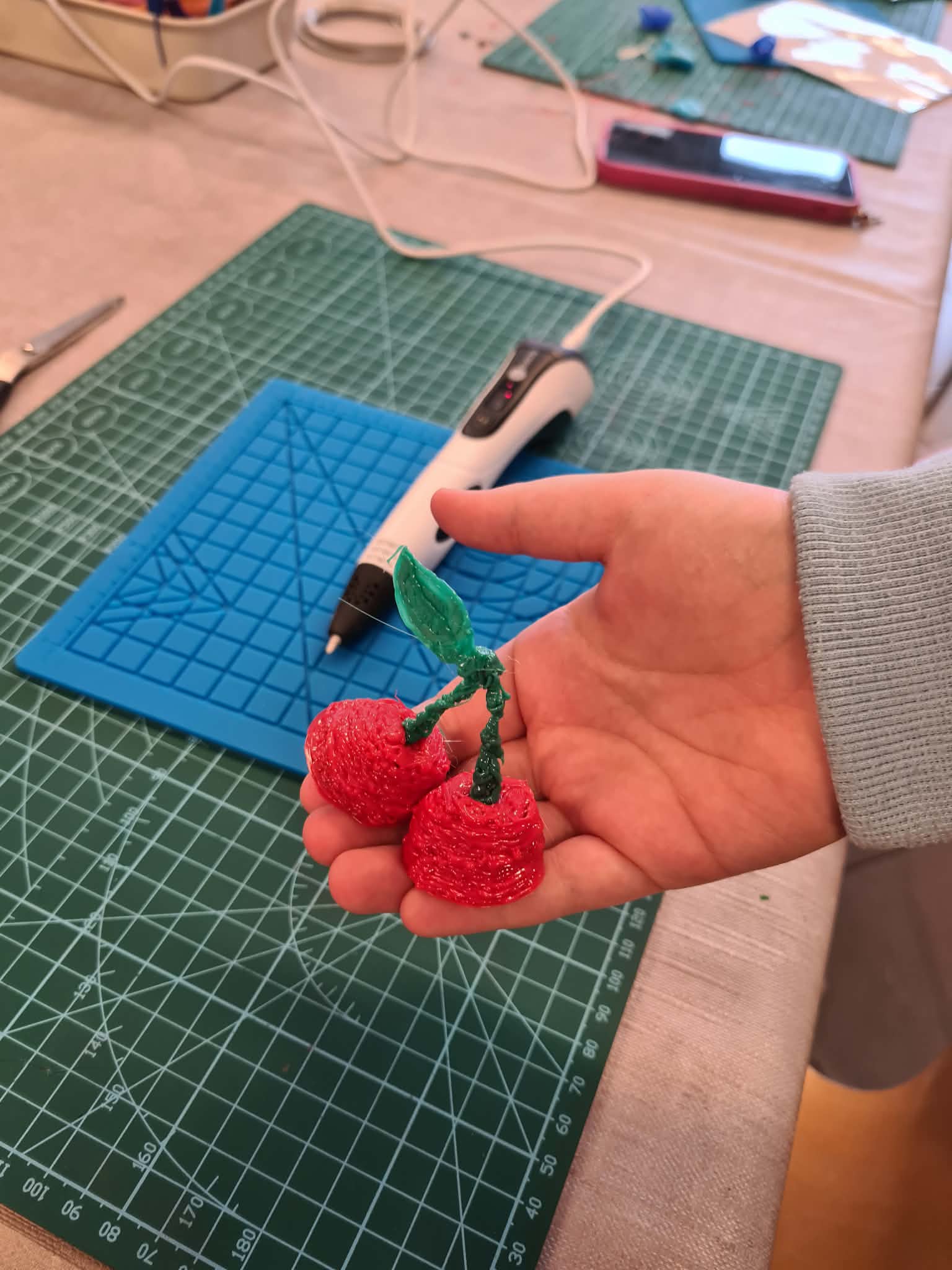 Dłoń trzyma czerwone wiśnie wykonane długopisem 3D na macie roboczej.