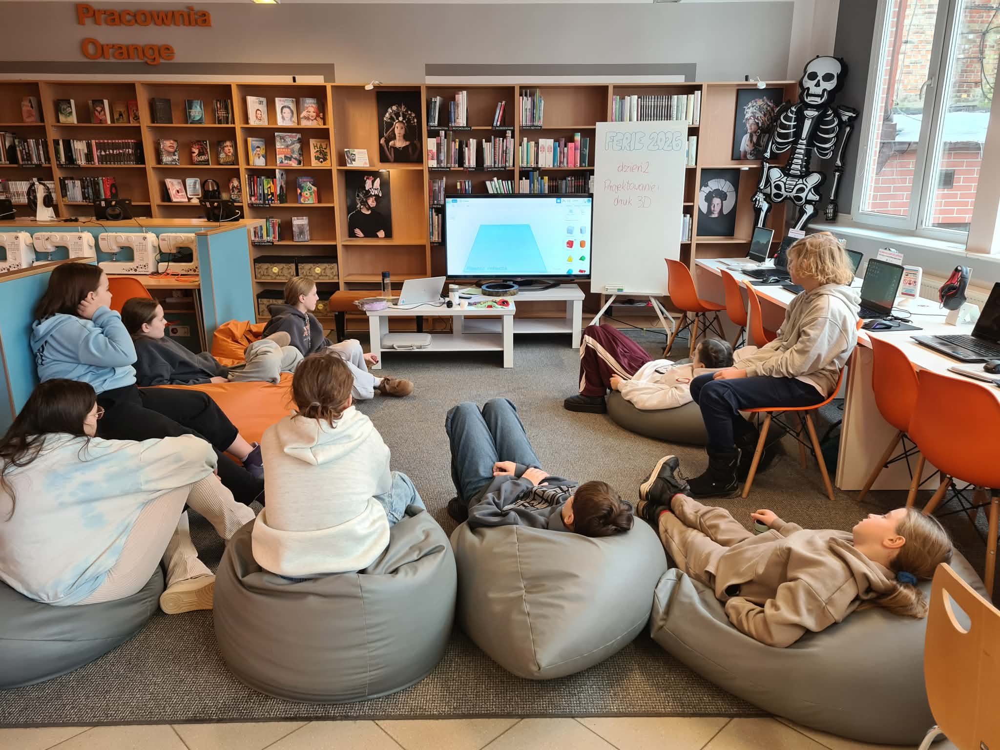 Grupa młodzieży siedzi na pufach i ogląda prezentację w bibliotece.