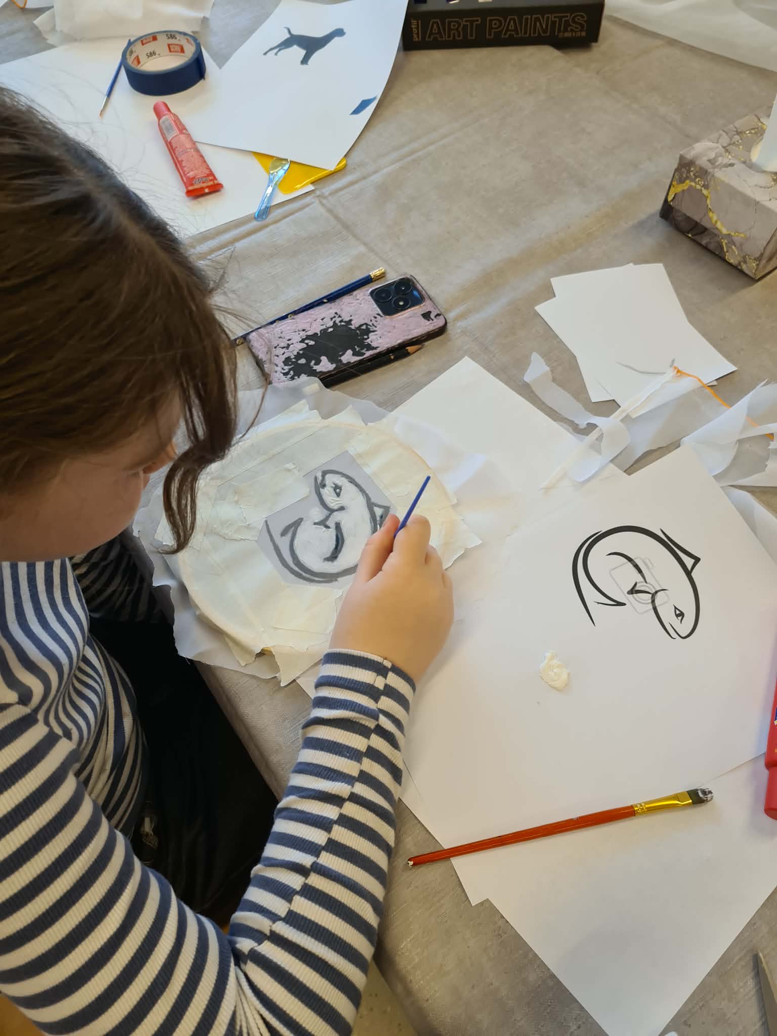 Dziecko maluje wzór na tkaninie, obok leżą kartki z rysunkami.