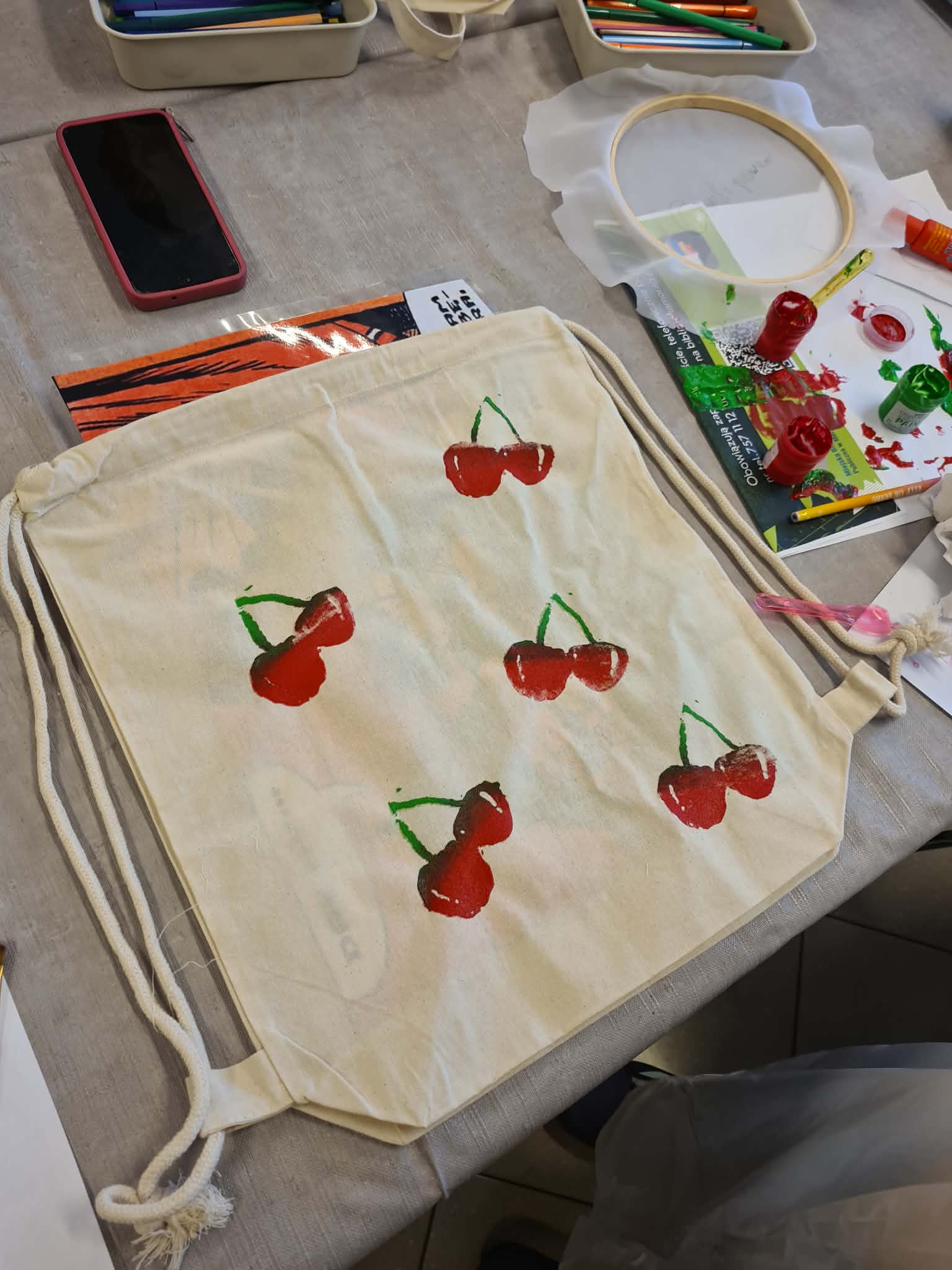 Płócienny worek z ręcznie malowanymi czerwonymi wiśniami, leży na stole z farbami i materiałami plastycznymi.
