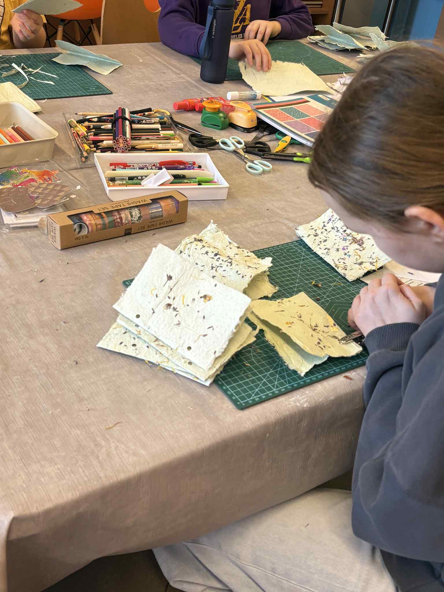 Stół warsztatowy z arkuszami papieru czerpanego, kredkami, nożyczkami i klejem.