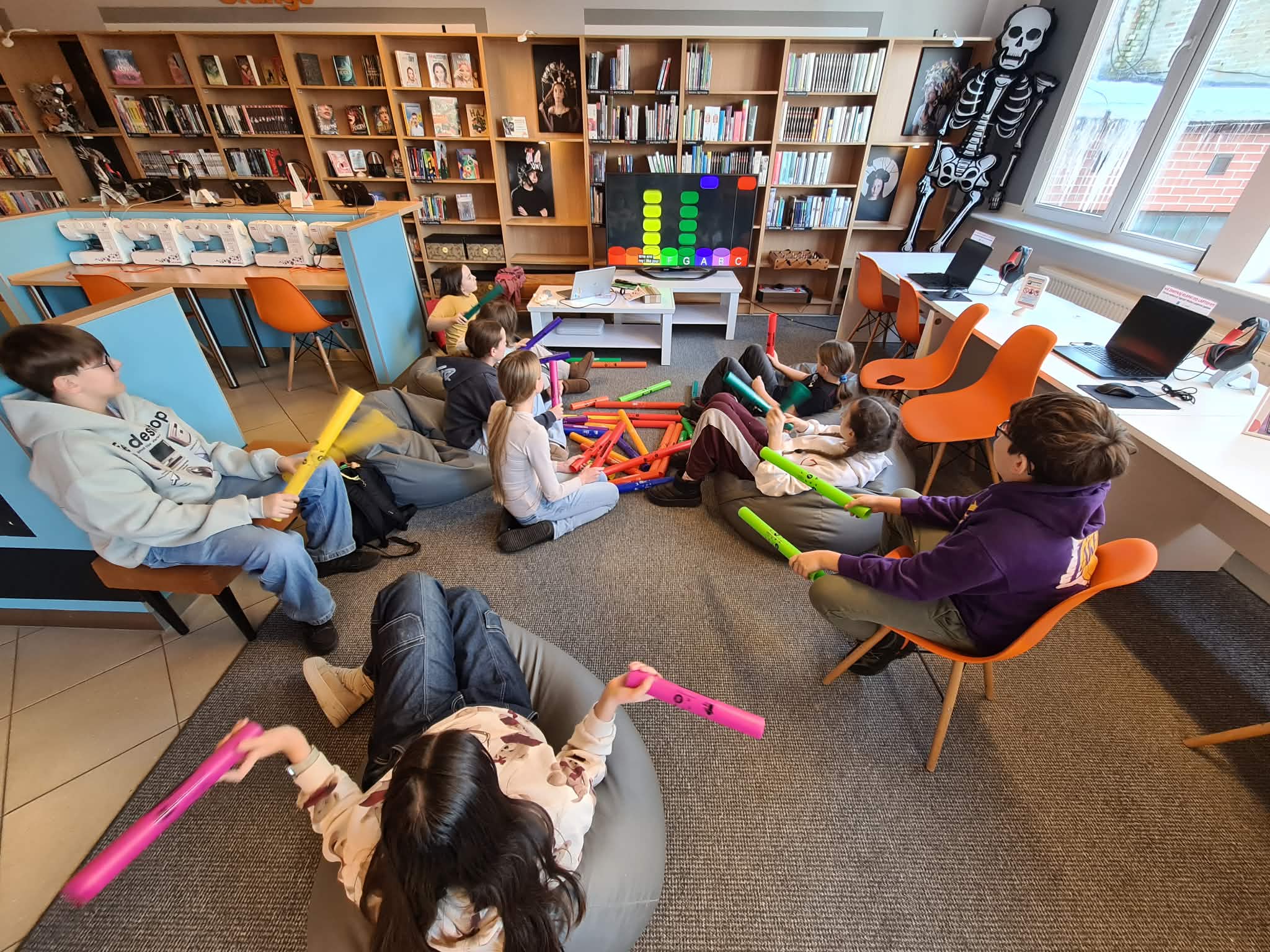 Dzieci siedzą na pufach i grają na kolorowych tubach muzycznych w bibliotece.