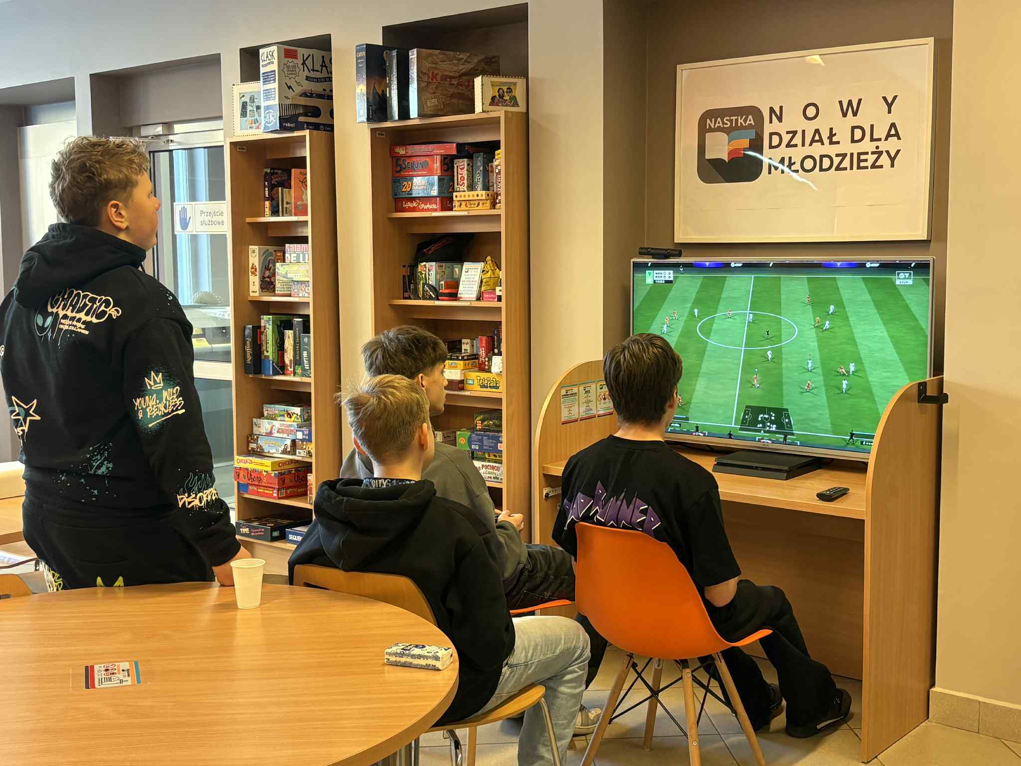 Nastolatkowie grają w grę piłkarską na dużym ekranie w bibliotece. Obok widoczne półki z grami planszowymi i książkami.